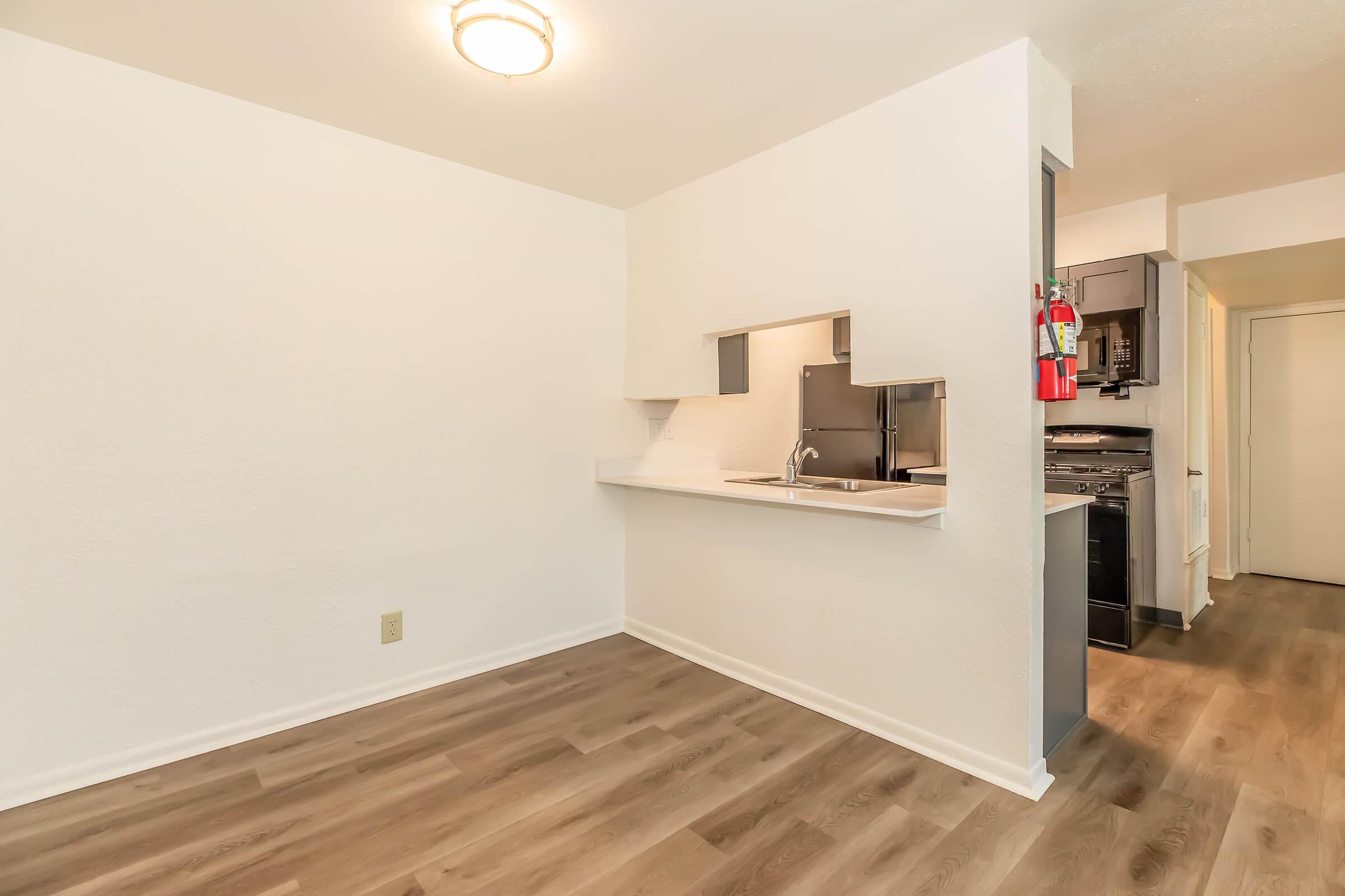 A small, modern kitchen with a white wall and light wooden flooring. The kitchen features a sink and a section for appliances. A doorway leads to another room, and there is a fire extinguisher mounted on the wall. The overall decor is minimal and contemporary, with ample natural light.