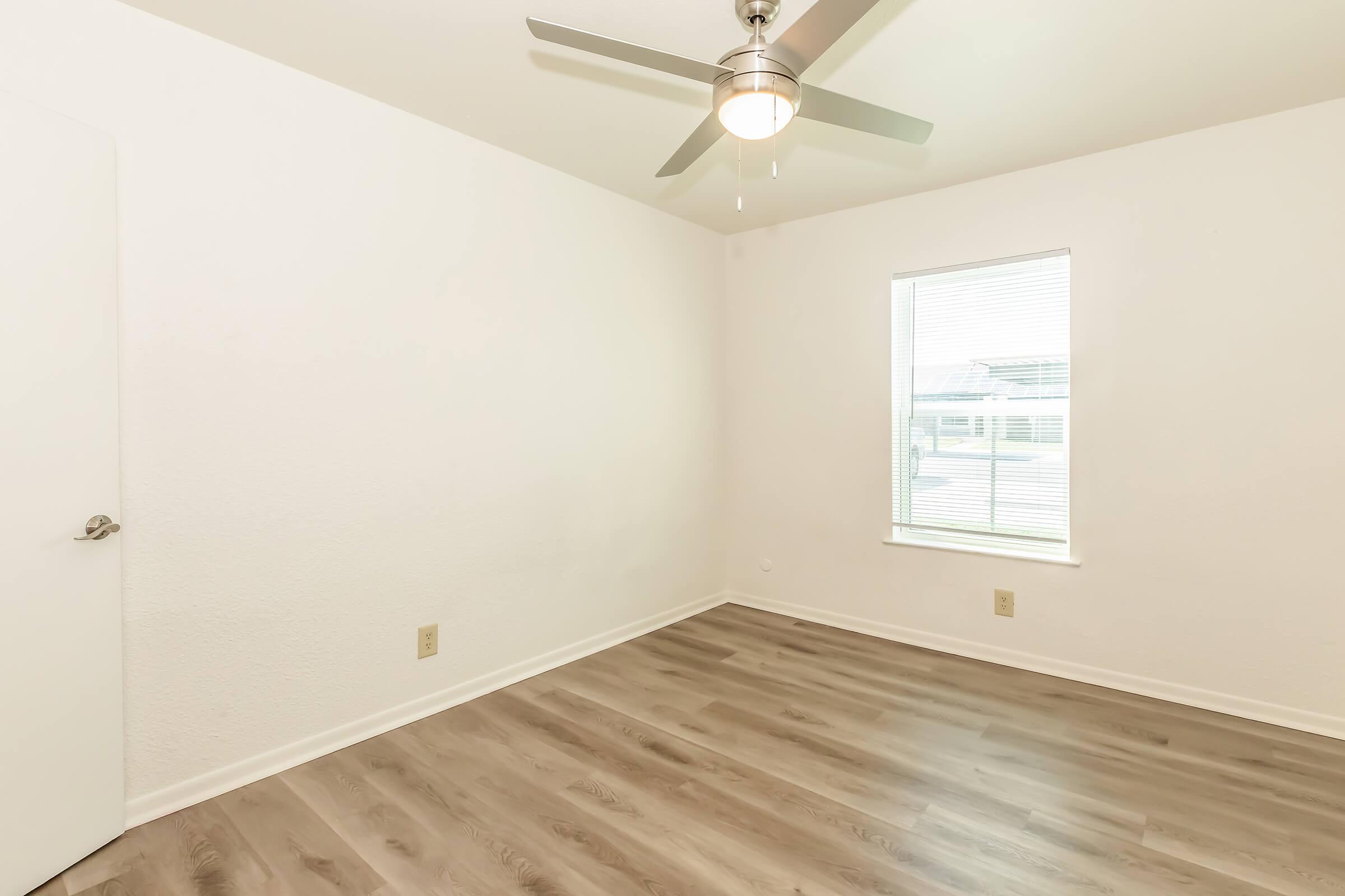 Empty room featuring light-colored walls, a ceiling fan with a metal finish, and a single window allowing natural light. The floor is covered with wood-like laminate, and there is a white door visible on the left.