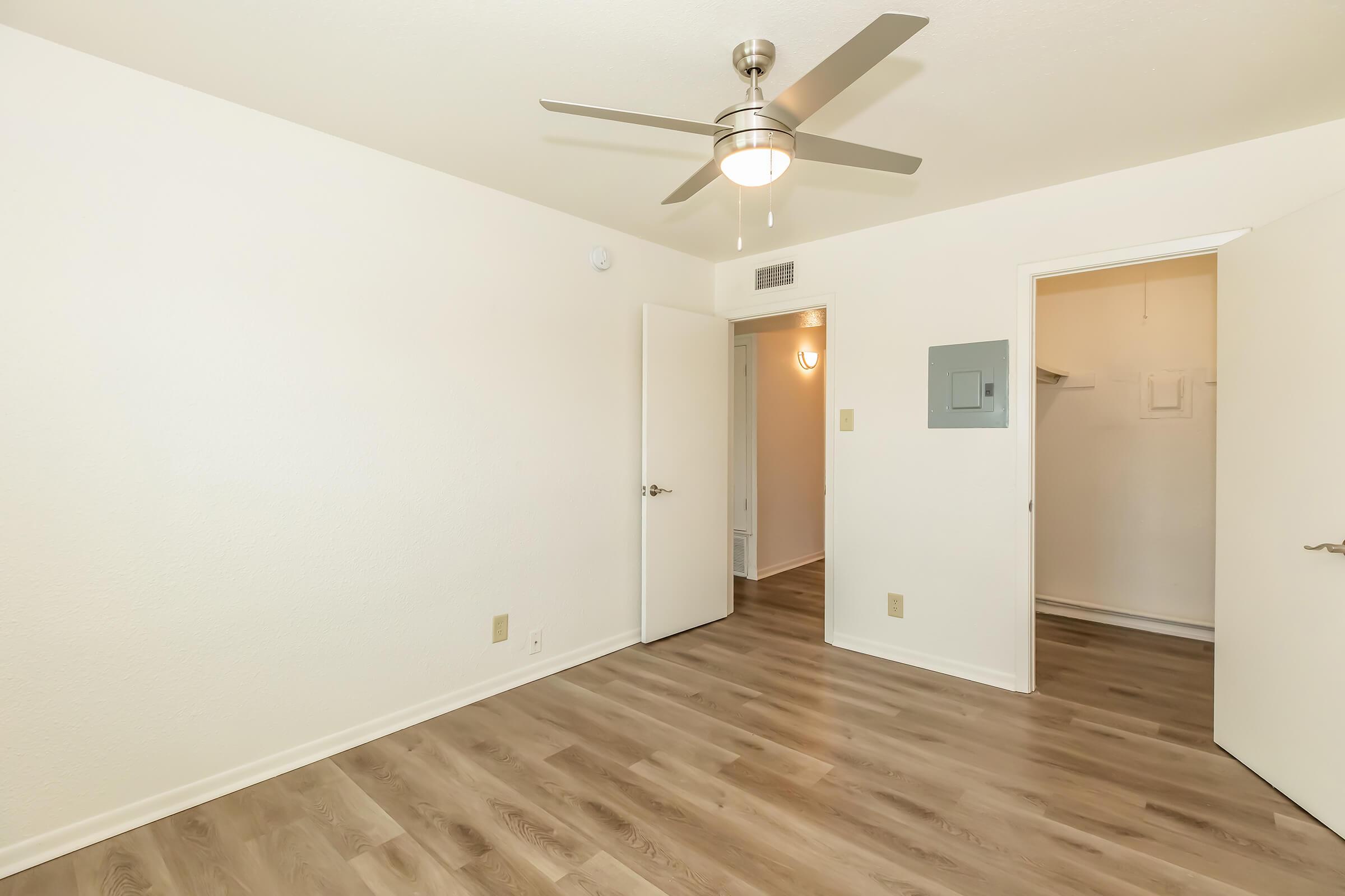 A bright, empty room featuring light-colored walls and wooden flooring. A ceiling fan is mounted in the center, and there are two doors: one leading to a closet and the other possibly to a bathroom or hallway. Natural light illuminates the space, creating a fresh and open atmosphere.