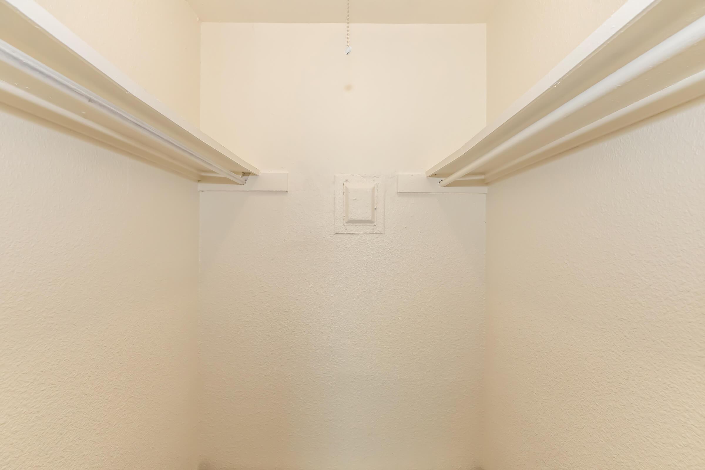 Empty closet with white walls and two shelves on either side. The space is devoid of any items, creating a clean and minimalist appearance. A small rectangular patch is visible on the back wall, suggesting a former fixture or vent. The lighting is bright and neutral.