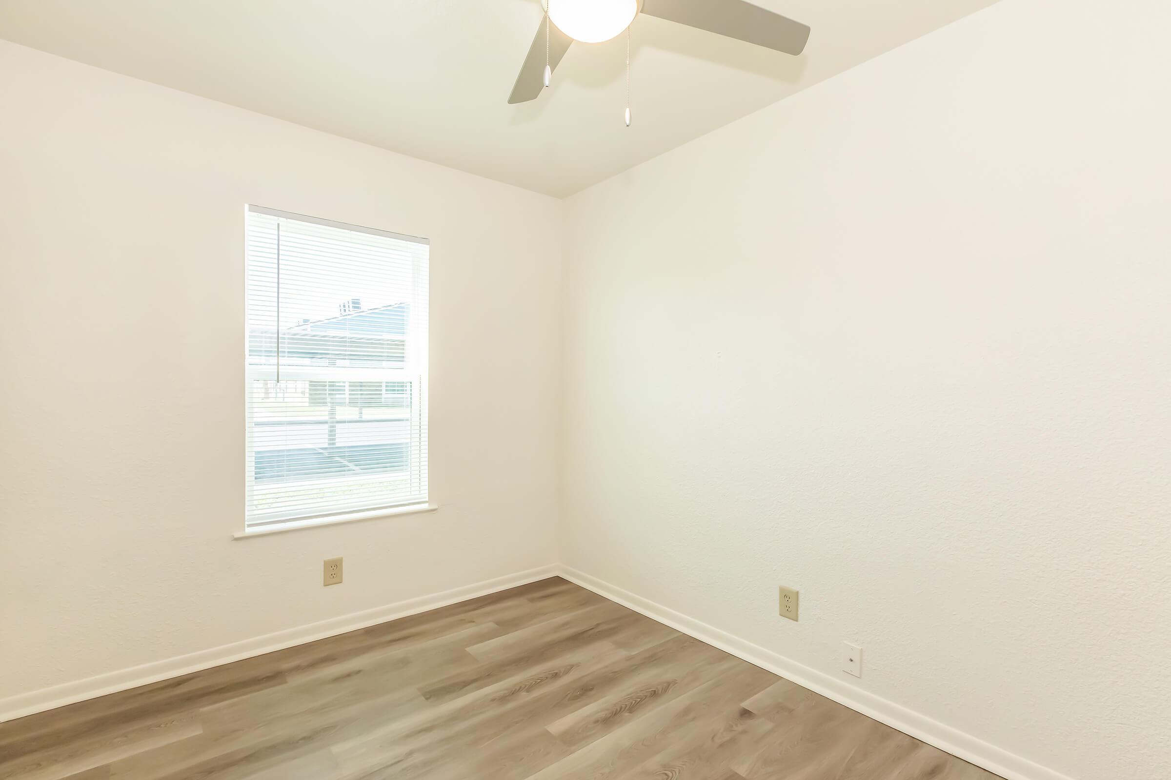 Empty room with light-colored walls and a ceiling fan. The floor is covered with wood-like laminate, and there is a window with blinds allowing natural light to enter. The overall space appears bright and modern, suitable for various uses.