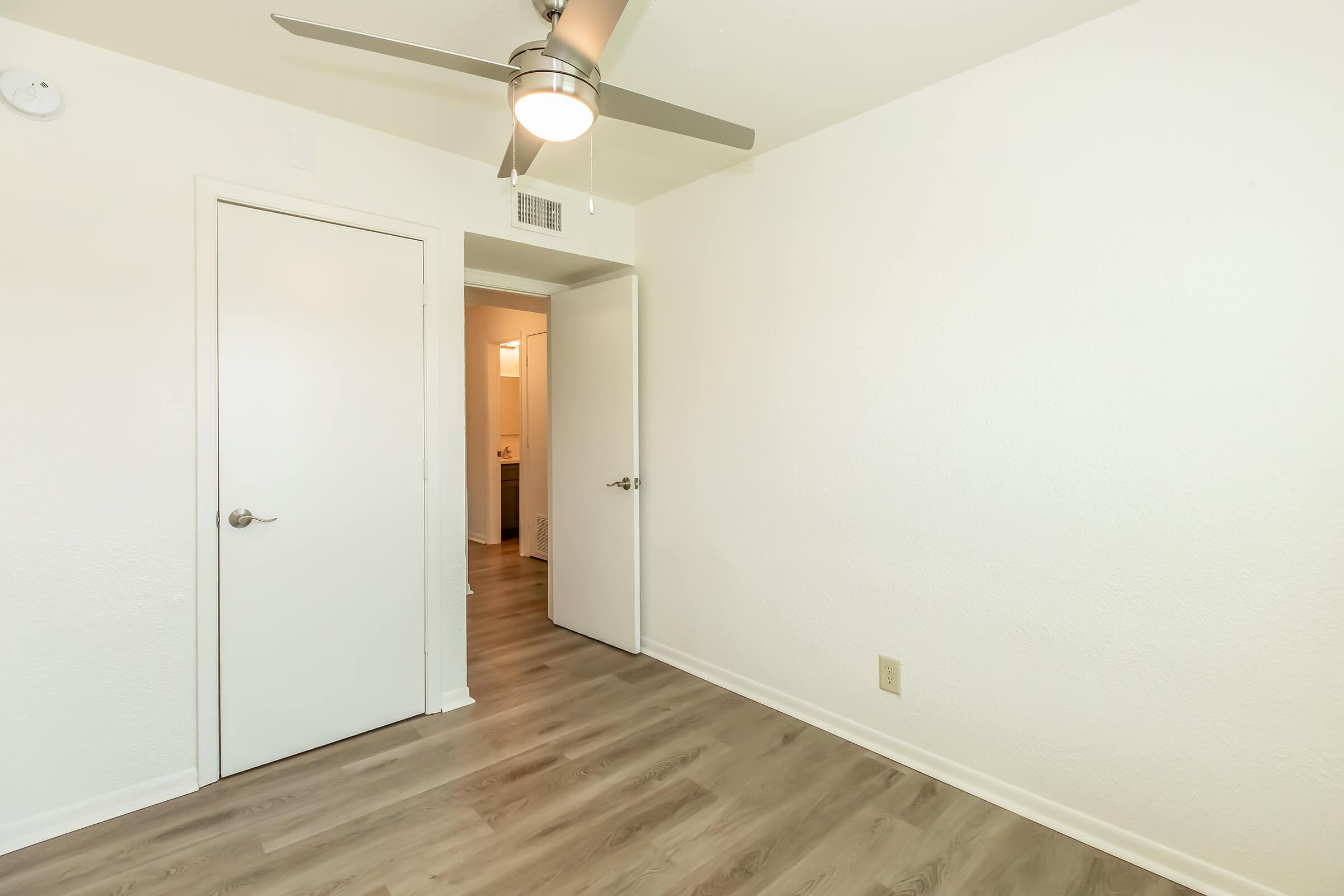 Empty room featuring a light-colored wall, a ceiling fan, and laminate flooring. A closed door on the left leads to another room, while an open door on the right shows a glimpse of a bathroom area. The space is well-lit and minimalistic, emphasizing a clean and modern aesthetic.