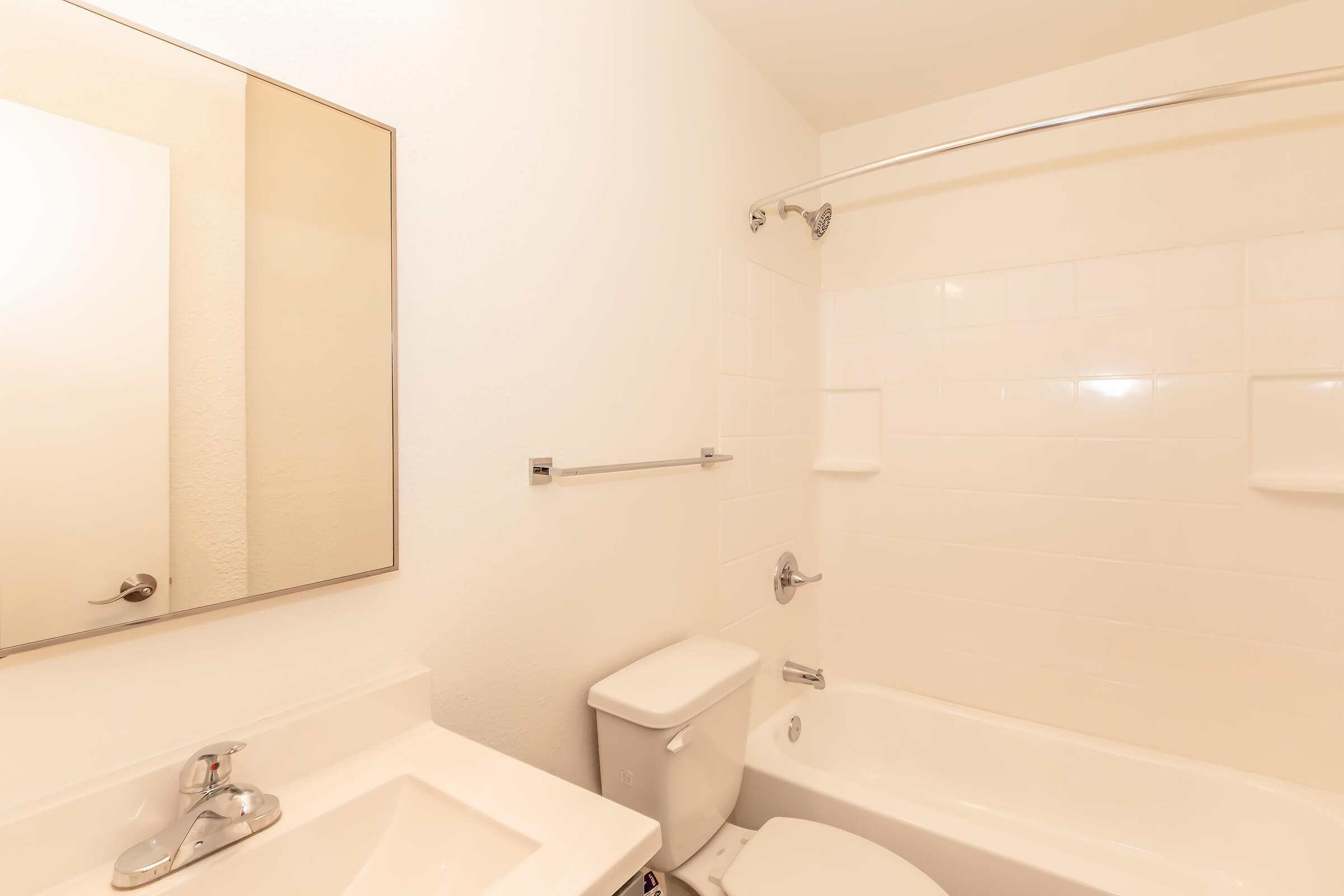A clean, minimalist bathroom featuring a white tiled bathtub with a shower, a rectangular mirror above a sink, and a white toilet. The walls and fixtures are bright and simple, creating a spacious and light atmosphere.