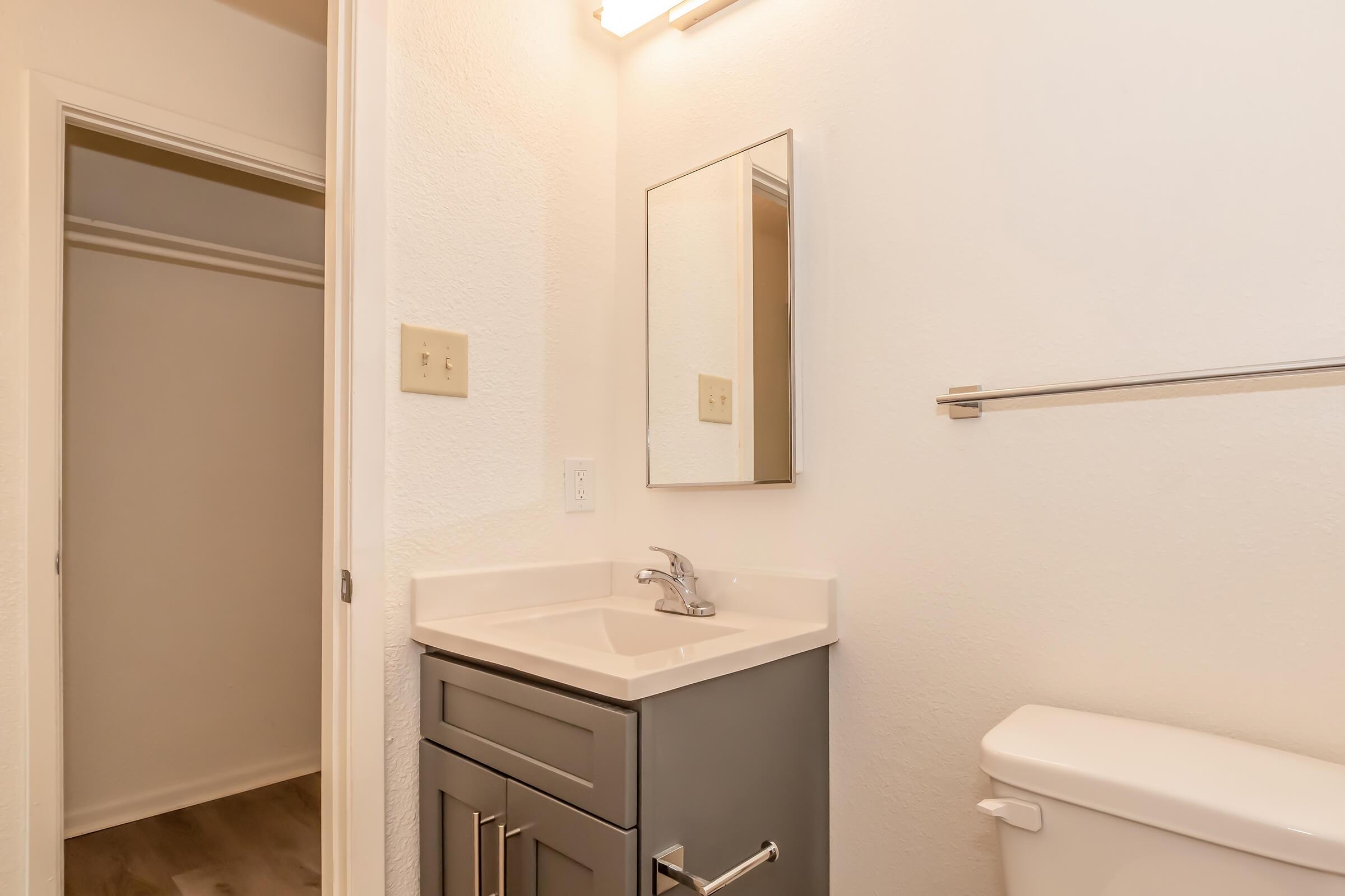 A clean and modern bathroom featuring a compact vanity with a sink, a mirror above it, a toilet on the right, and a doorway leading to a closet. The walls are painted white, and the floor has a light wood finish, creating a bright and inviting atmosphere.