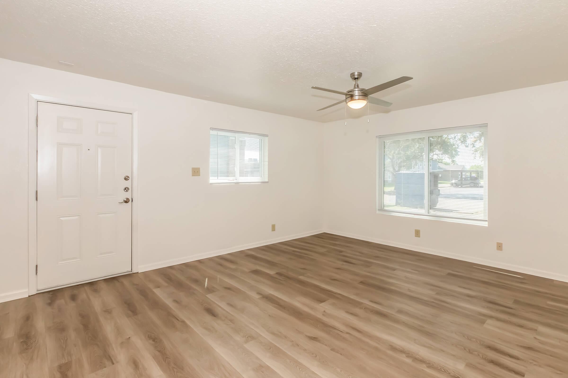 Bright and airy living room featuring a white wall, light wood flooring, and a ceiling fan. Large windows allow natural light to flood the space, and the entrance door is visible on the left side. The room is empty, providing a fresh and neutral backdrop suitable for decorating.