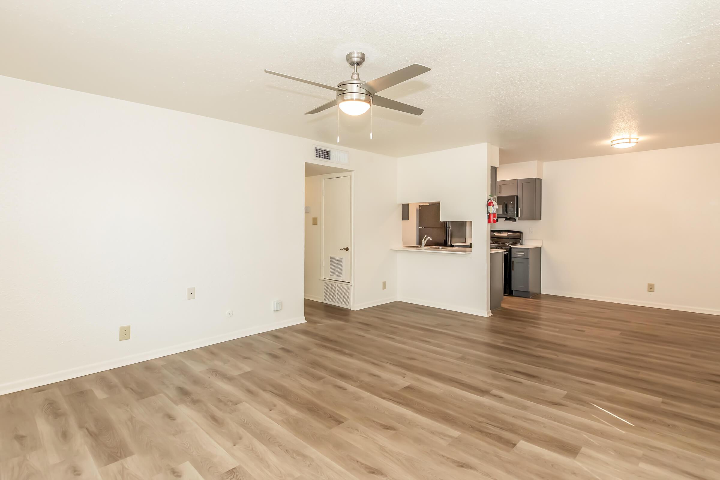 Spacious and modern living area with light-colored walls and laminate flooring. A ceiling fan provides airflow, and a kitchen is visible in the background with dark cabinets and appliances. The room features a neutral color palette, creating a bright, inviting atmosphere.