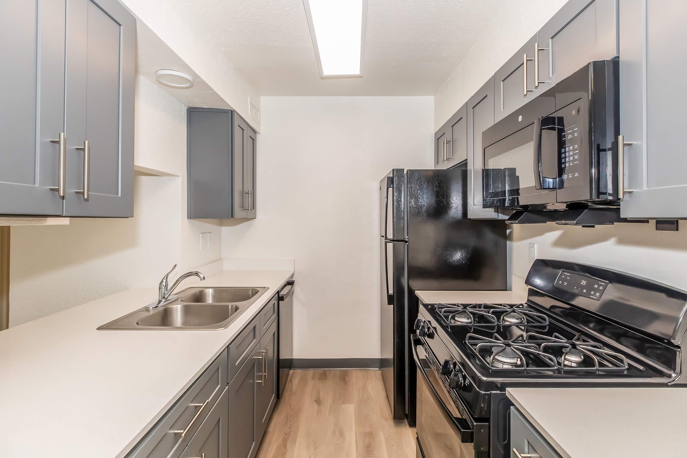 Modern kitchen featuring dark gray cabinetry, stainless steel appliances including a gas stove and microwave, double sink, and a light-colored countertop. The space is well-lit with overhead lighting and has a clean, minimalist design.