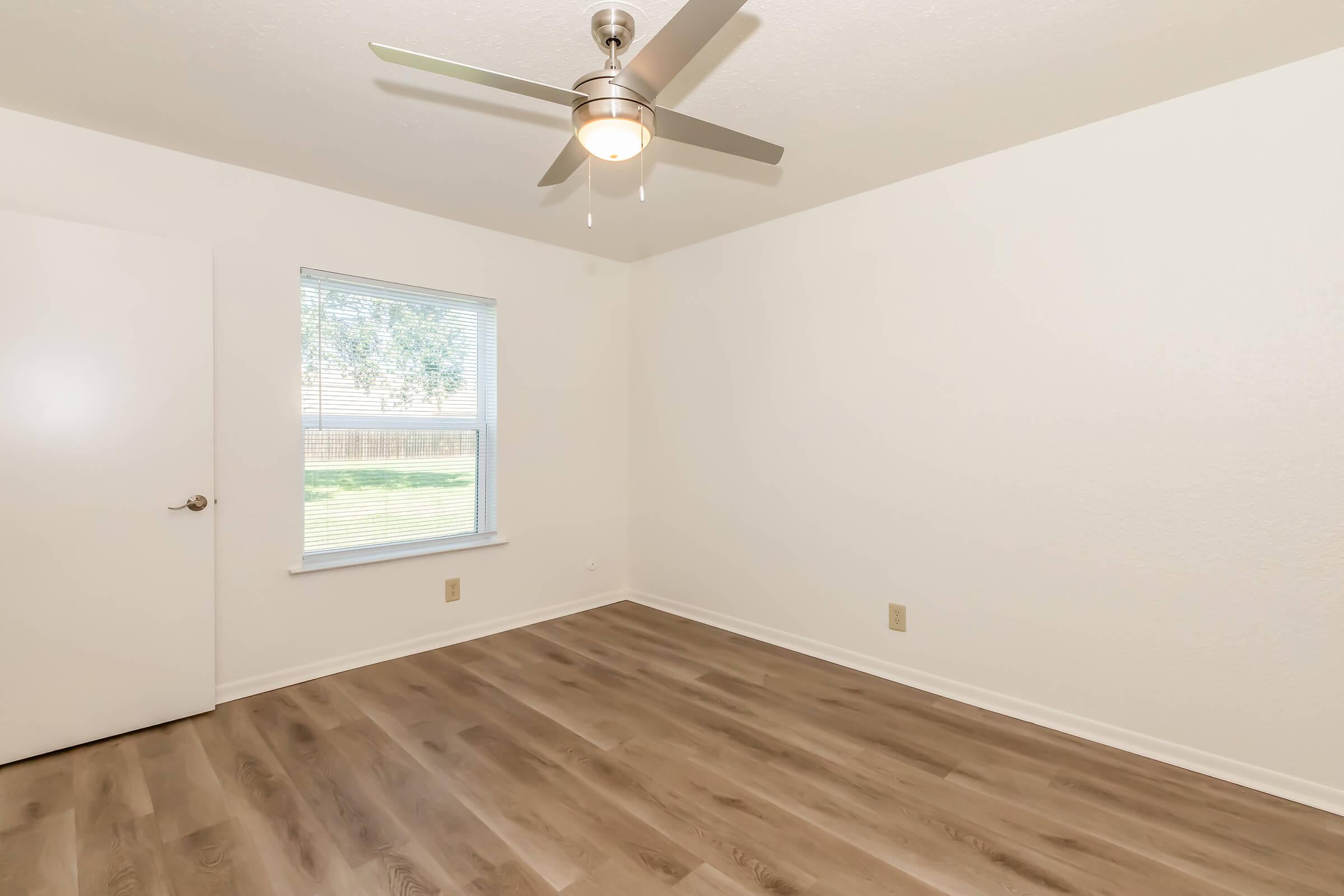 A clean, empty room with light-colored walls and a wood-like laminate floor. A ceiling fan is installed, and there is a window with blinds that lets in natural light, offering a view of greenery outside. A white door is partially visible on the left side of the image.