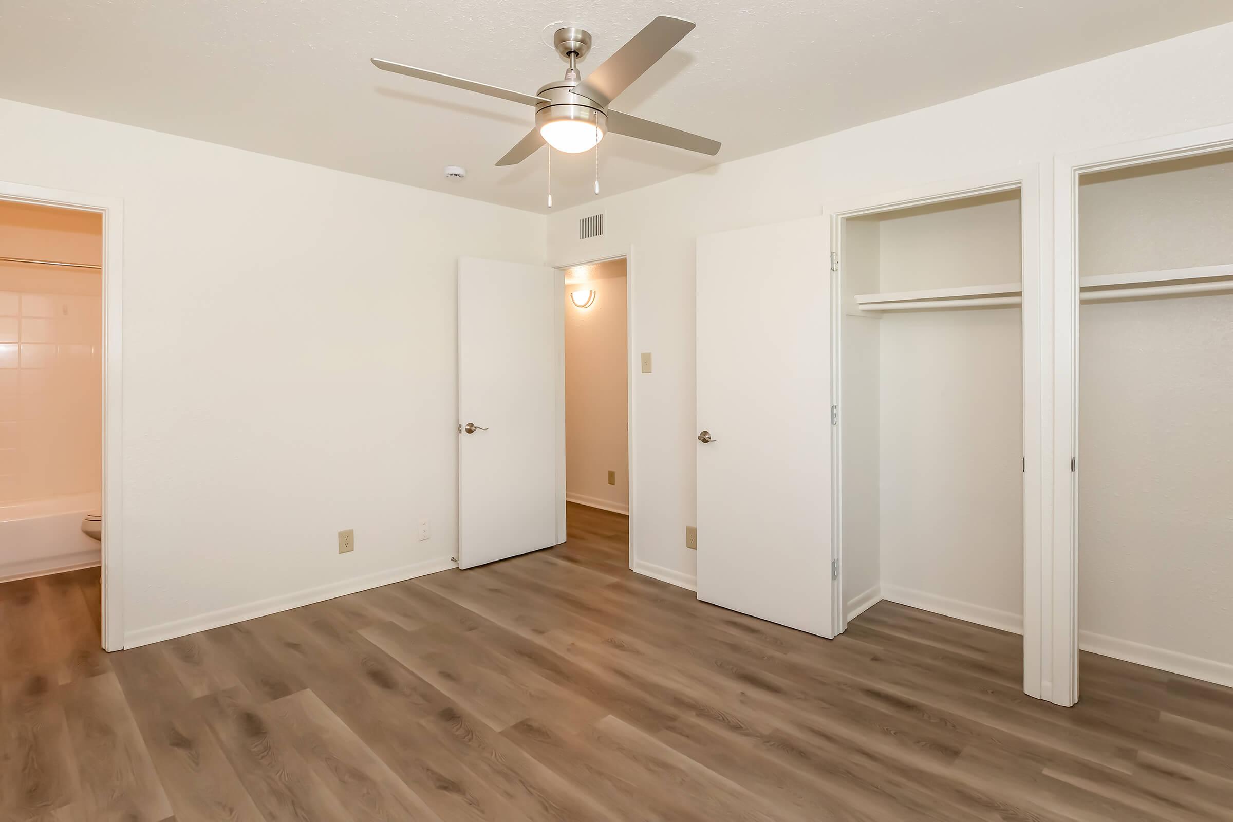 A spacious room featuring light-colored walls and hardwood-like flooring. There are two white doors leading to closets on the right, and a ceiling fan overhead. A bathroom with a tub is visible through an open door at the back. Natural light brightens the space, creating an inviting atmosphere.