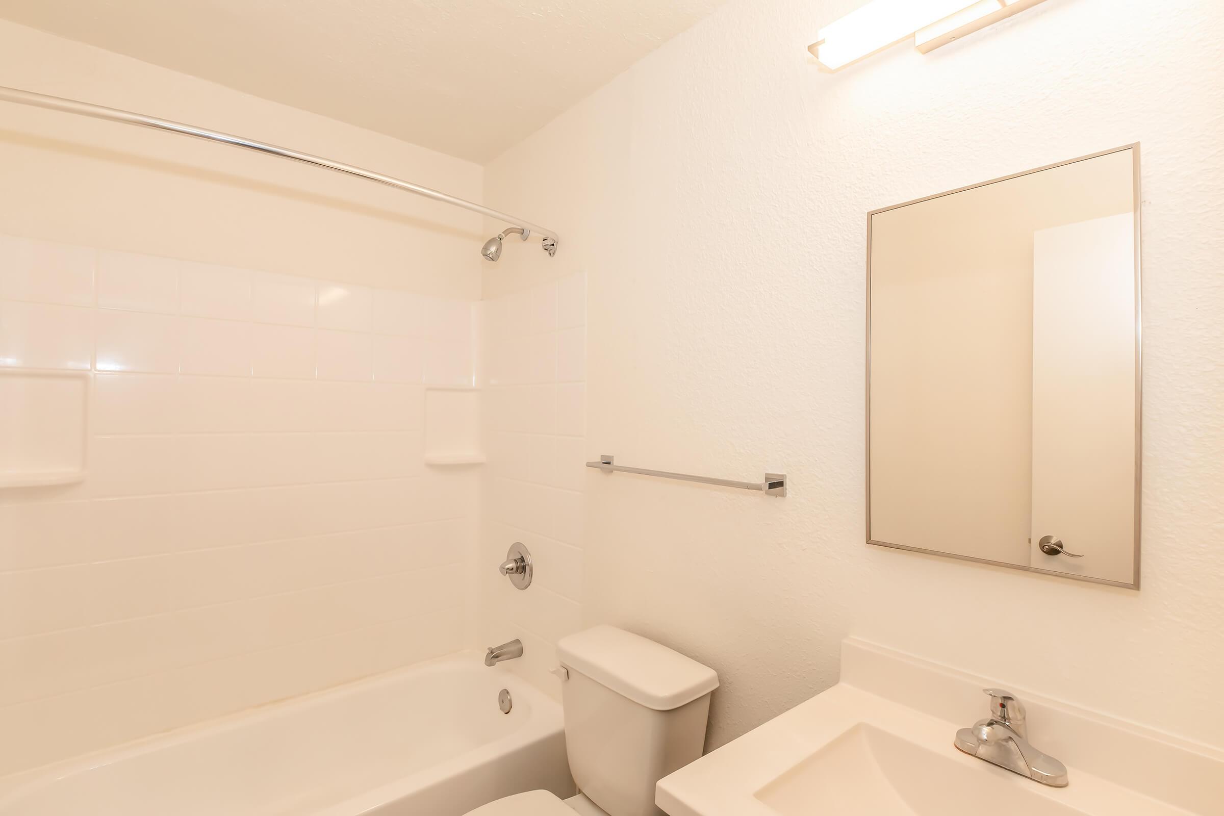 A clean, minimalistic bathroom featuring a bathtub with an attached shower, a toilet, and a sink with a mirror above it. The walls are white with white tiles, giving the space a bright and simple appearance. Light fixtures are mounted on the wall above the mirror.