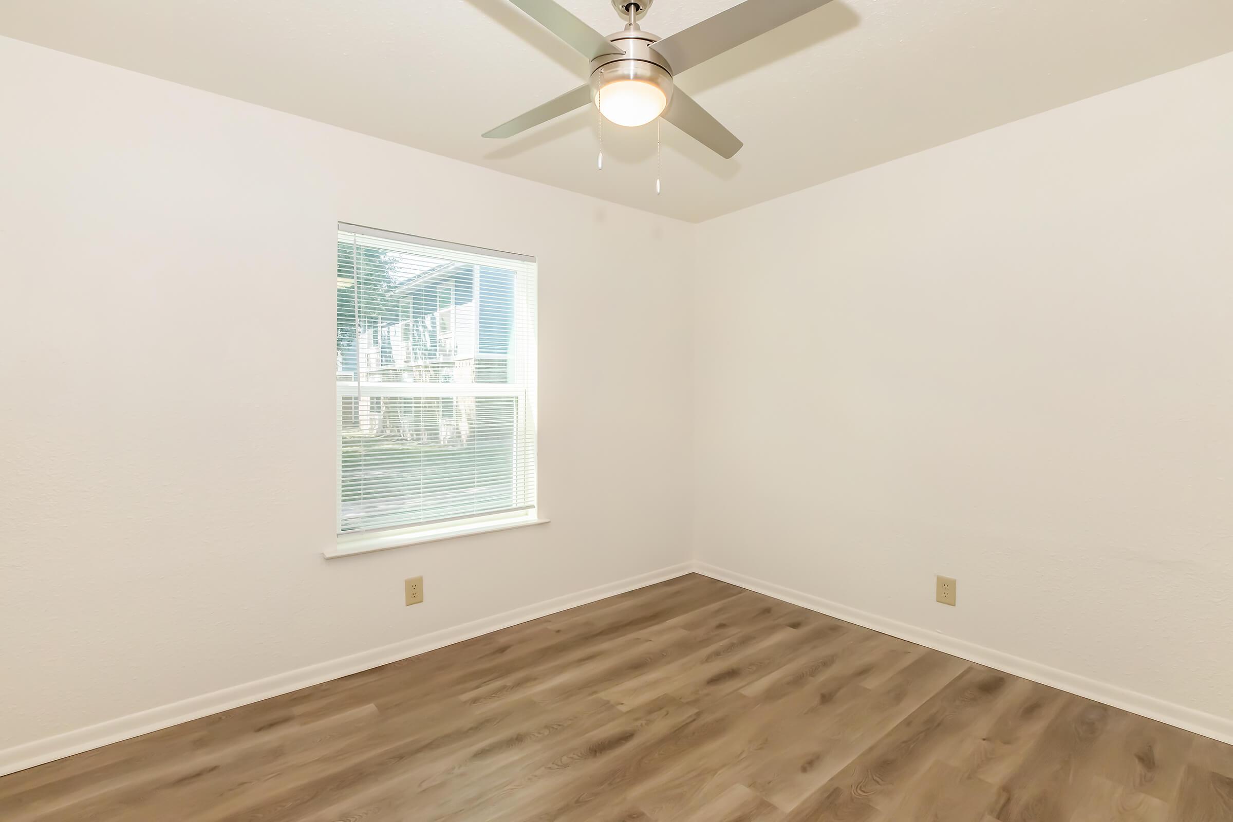 Empty room with light-colored walls and hardwood flooring. A ceiling fan is mounted on the ceiling, and there is a window allowing natural light to enter. The space appears clean and uncluttered, ready for furniture placement.
