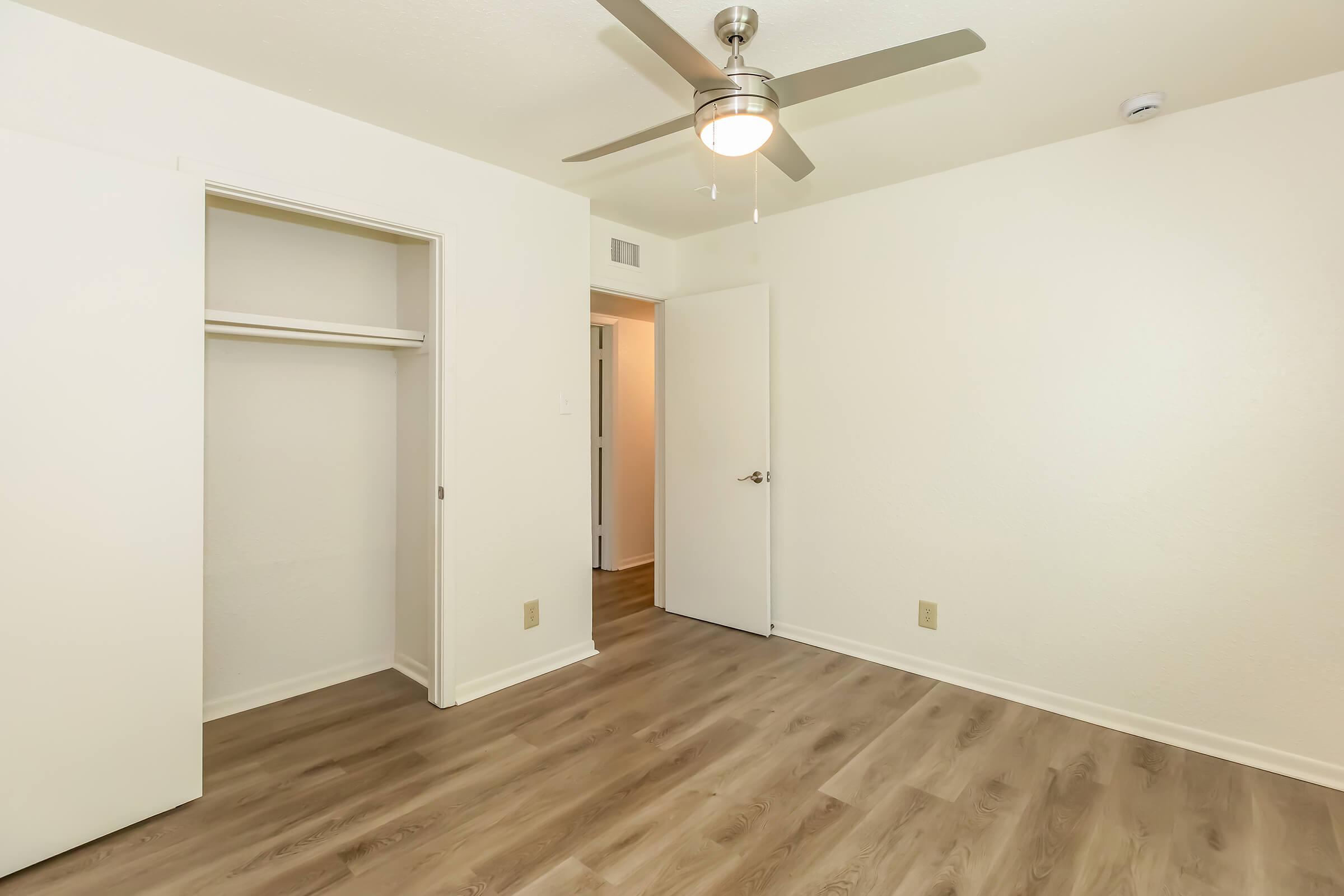 A small, well-lit room with light-colored walls and wooden flooring. It features a ceiling fan, a closet with sliding doors, and a doorway leading to another room. The space is empty and ready for furnishing, creating a clean and inviting atmosphere.