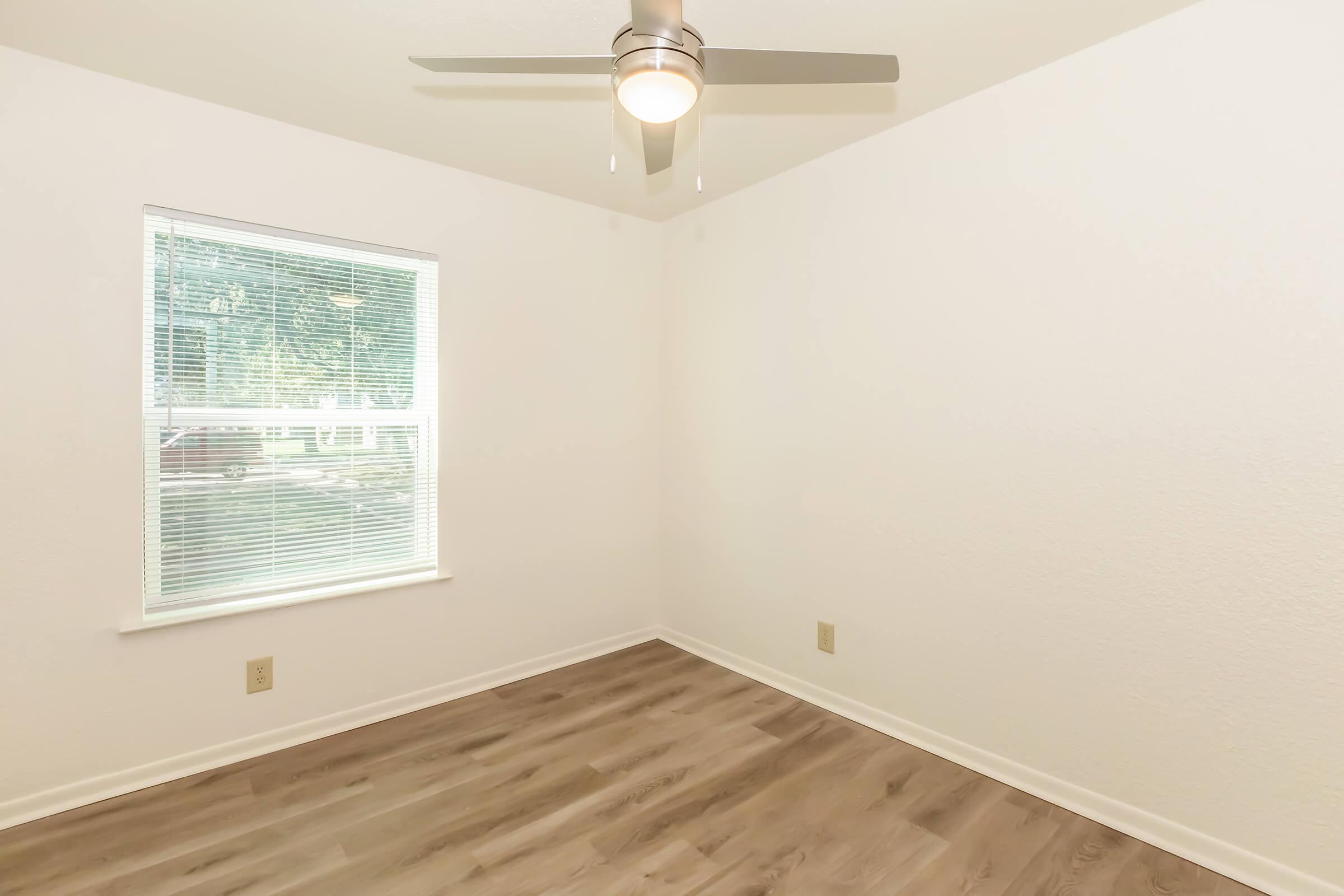 Empty room with light-colored walls and a light wooden floor. A large window allows natural light to enter, featuring a ceiling fan with four blades. The space is uncluttered and ready for furnishings, providing a bright and airy atmosphere.