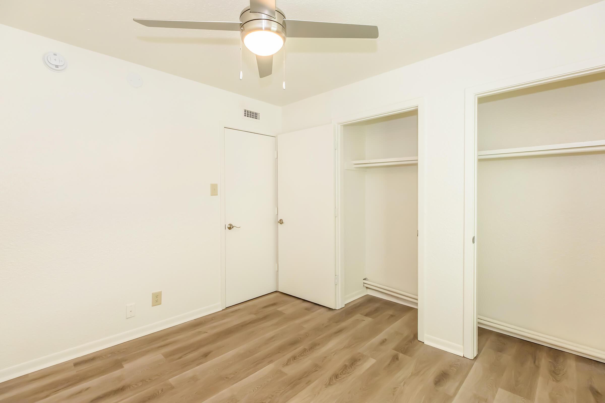 A clean, empty room featuring a light-colored wall, a ceiling fan, and two closet spaces. The floor is a light wood laminate. A door is visible on the left, and the overall space is well-lit and minimalistic, ideal for furnishing or decorating.