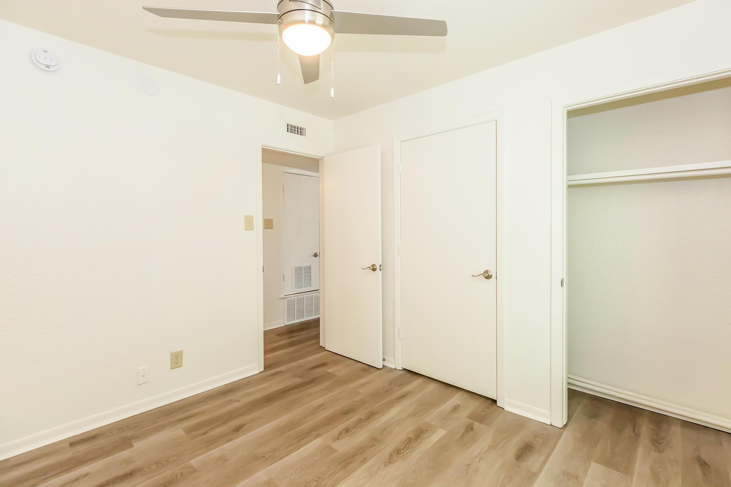 A simple interior of a room featuring light-colored walls, a ceiling fan with a light fixture, and a wooden floor. There are two doors, one leading to a closet and the other to another room. The space is well-lit and uncluttered, giving a sense of openness.