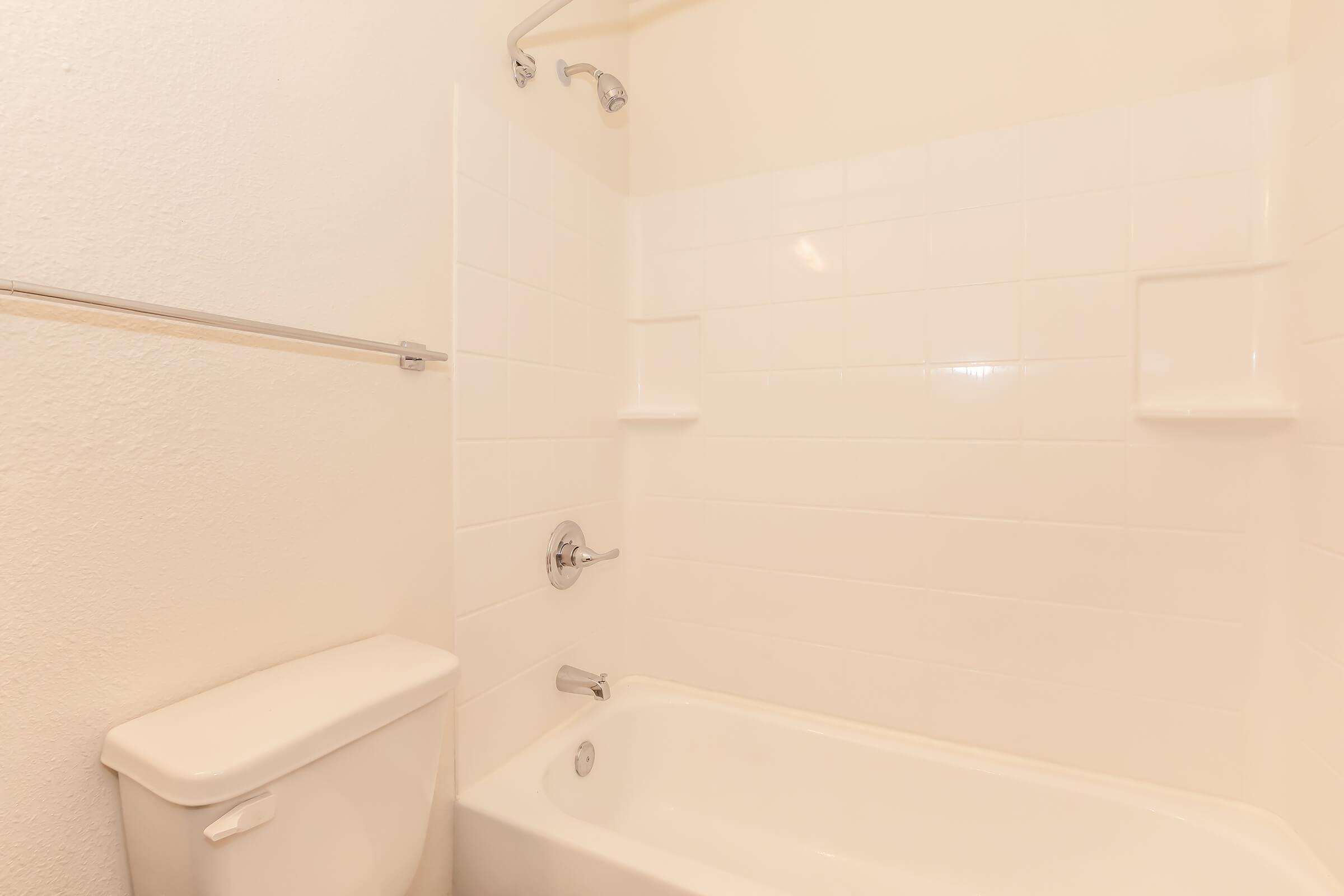 A clean, minimalistic bathroom featuring a white bathtub, a wall-mounted showerhead, and a toilet. The walls are tiled in light colors, creating a bright and spacious feel. There are built-in shelves in the wall for storage.