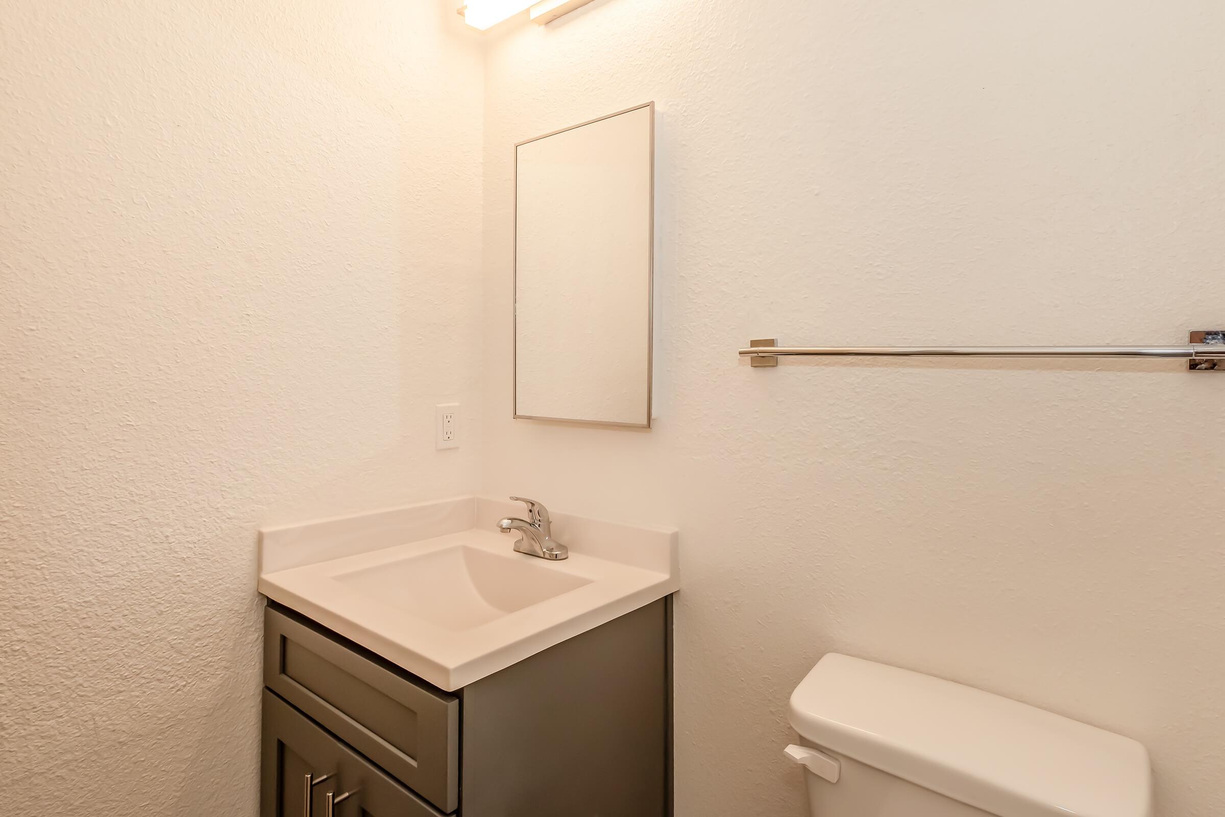 A clean, minimalist bathroom featuring a small vanity with a sink and a mirror above it. The walls are painted in a light color, and there's a toilet visible on the right. A towel bar is mounted on the wall, adding functionality to the space. The lighting is bright and welcoming.