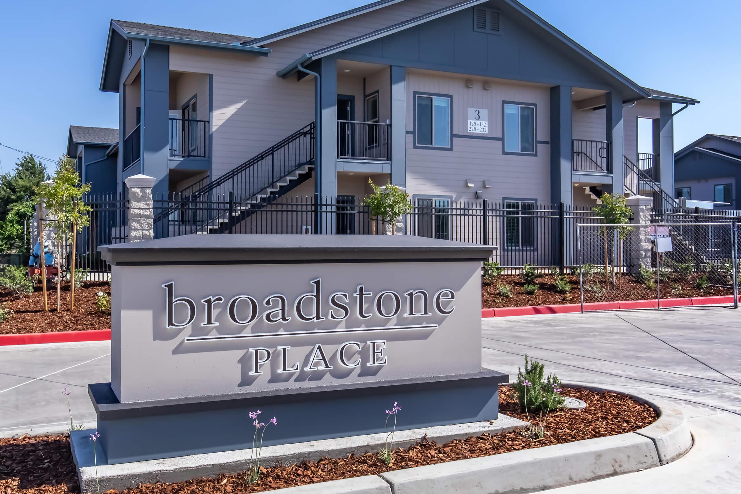 Sign for Broadstone Place in front of a modern apartment complex, featuring a two-story building with balconies. The landscaping includes small plants and mulch, with a clear blue sky in the background. The sign is prominent and displayed in a landscaped area near the entrance.
