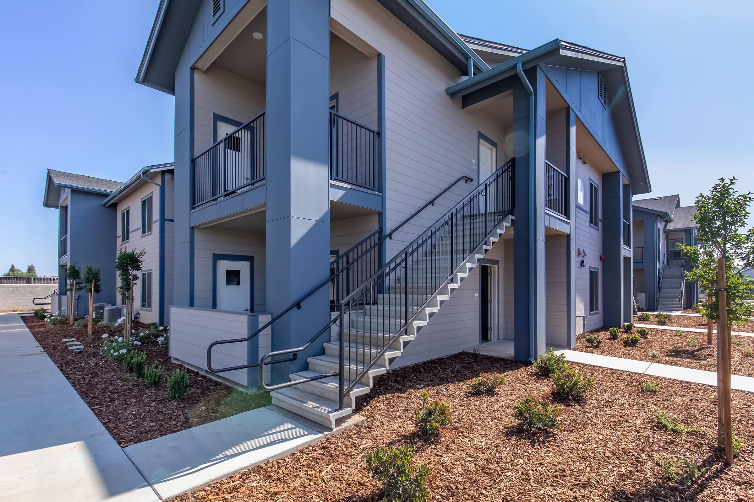 A modern apartment complex with light gray and blue exterior walls. The two-story building features a staircase leading to the upper level. Neatly landscaped beds with shrubs and flowers line the path, providing a welcoming appearance. Bright sunlight enhances the clarity of the scene.