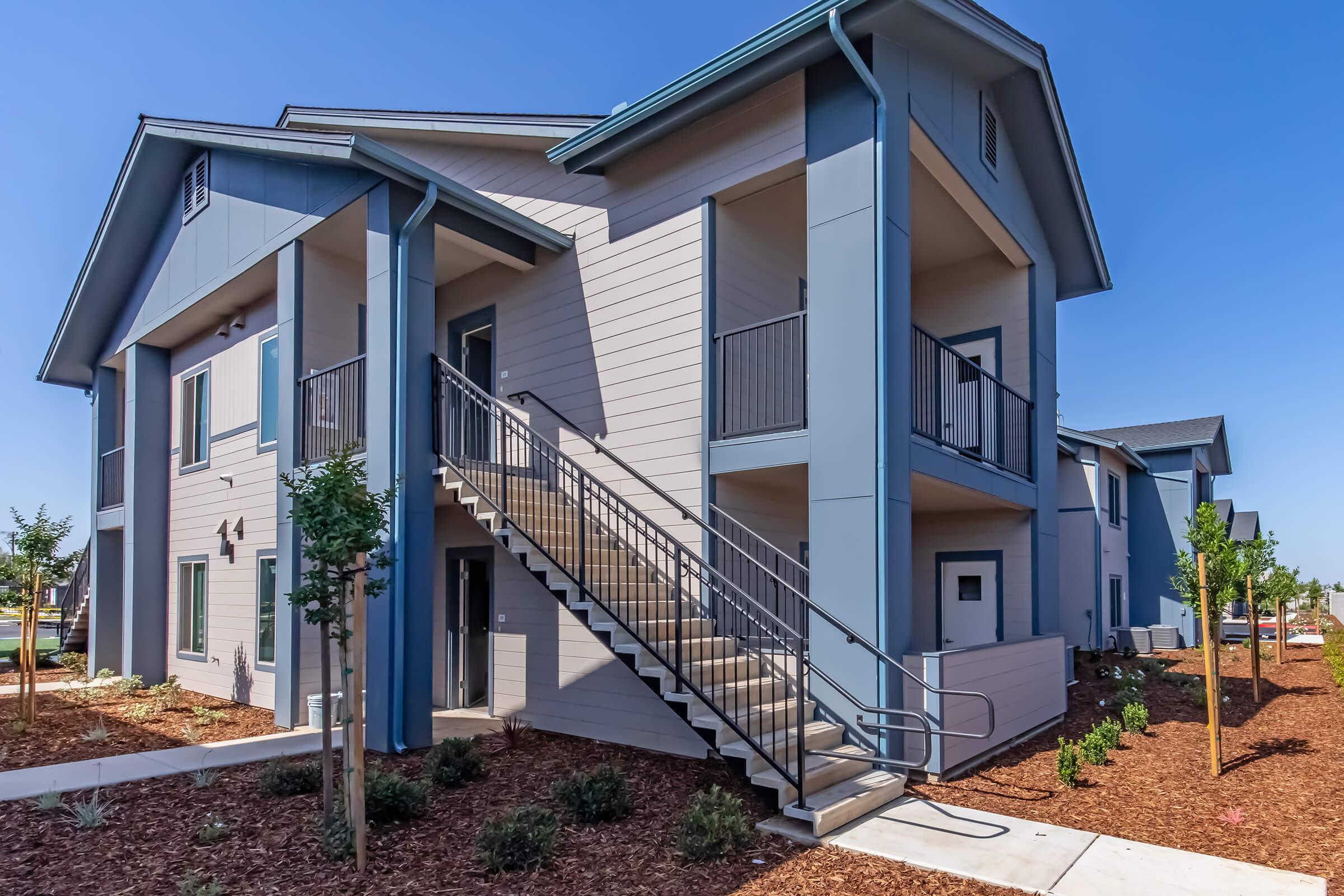 A modern two-story apartment building with a light blue exterior, featuring a staircase leading to the second floor. The surrounding area is landscaped with small trees and shrubs, under a clear blue sky.