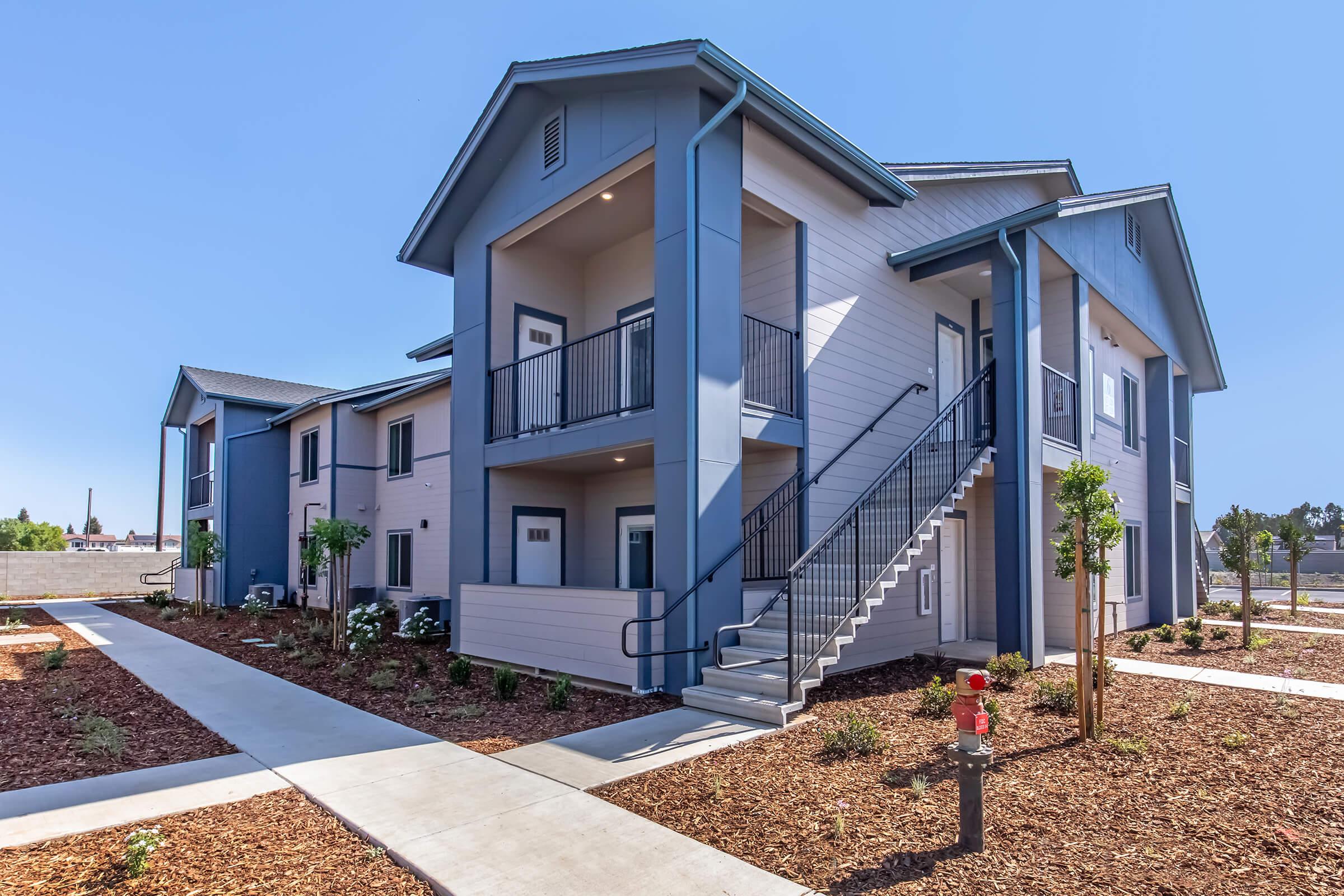 A modern two-story apartment building with blue and beige exteriors. The building features a staircase leading to the upper level and surrounded by landscaped grounds with mulch and small trees. Clear blue sky indicates sunny weather.