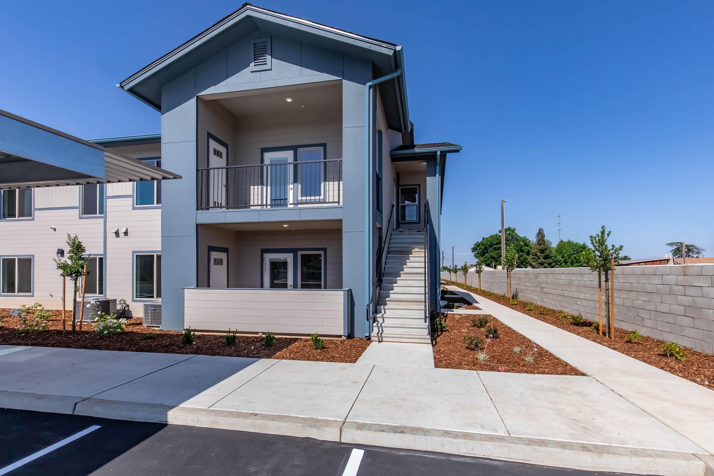 A modern two-story residential building with a balcony on the upper level. The exterior features a light blue color, surrounded by neatly landscaped areas with small trees and shrubs. There is a concrete pathway leading to the entrance, and parking spaces are visible in the foreground under a carport.