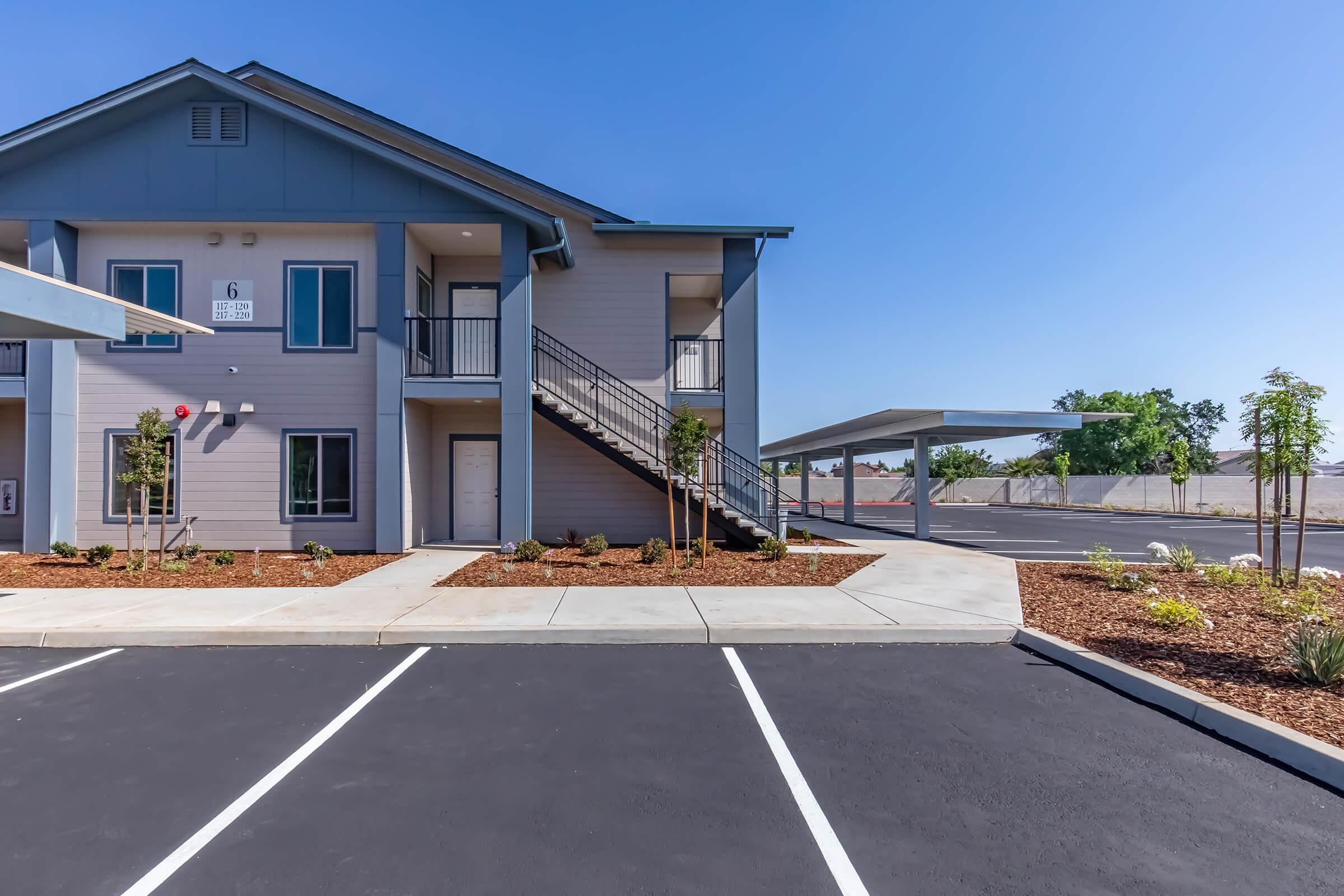 Exterior view of a two-story apartment building with a staircase, surrounded by landscaped areas with small plants and mulch. In the foreground, a newly paved parking lot with marked parking spaces and covered carports is visible. The sky is clear and blue.