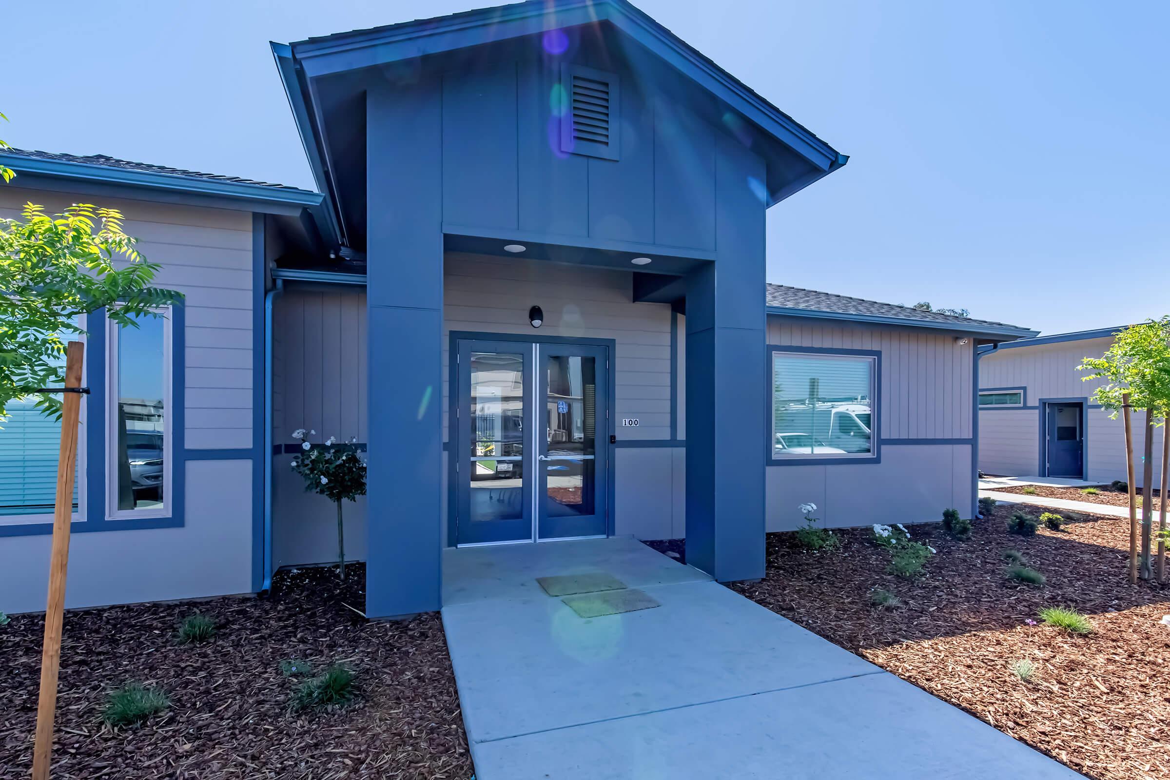 A modern building entrance featuring a blue and gray exterior. The front path is lined with small plants, and there are large windows on either side. The entrance is highlighted by a covered porch and a glass door, providing a welcoming atmosphere.