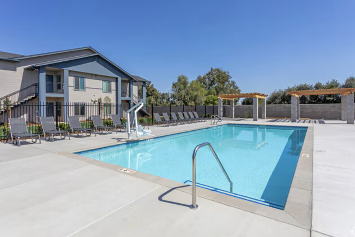 A clear blue swimming pool surrounded by lounge chairs and shaded seating areas. The poolside features a ladder for easy access, and a modern building can be seen in the background under a bright blue sky. The area is well-maintained with greenery around the pool.