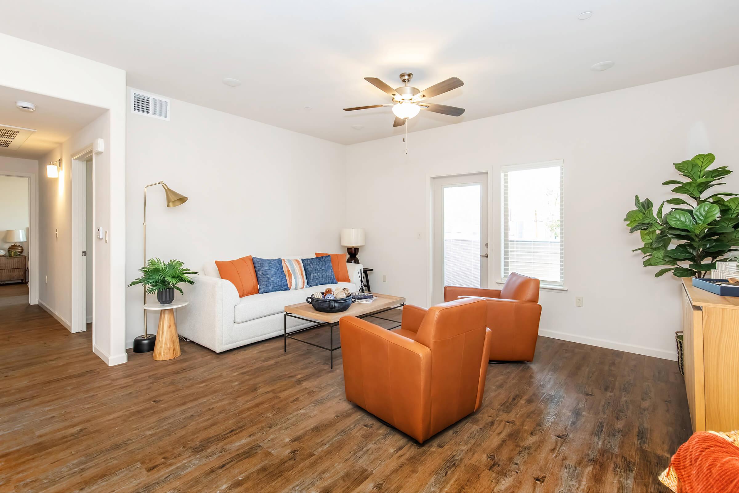 A modern living room featuring a white sofa with blue and orange accent pillows, two orange armchairs, a coffee table with decorative items, and a large plant. Natural lighting comes from large windows, and there is a ceiling fan above. The flooring is a warm wood finish, adding a cozy feel to the space.