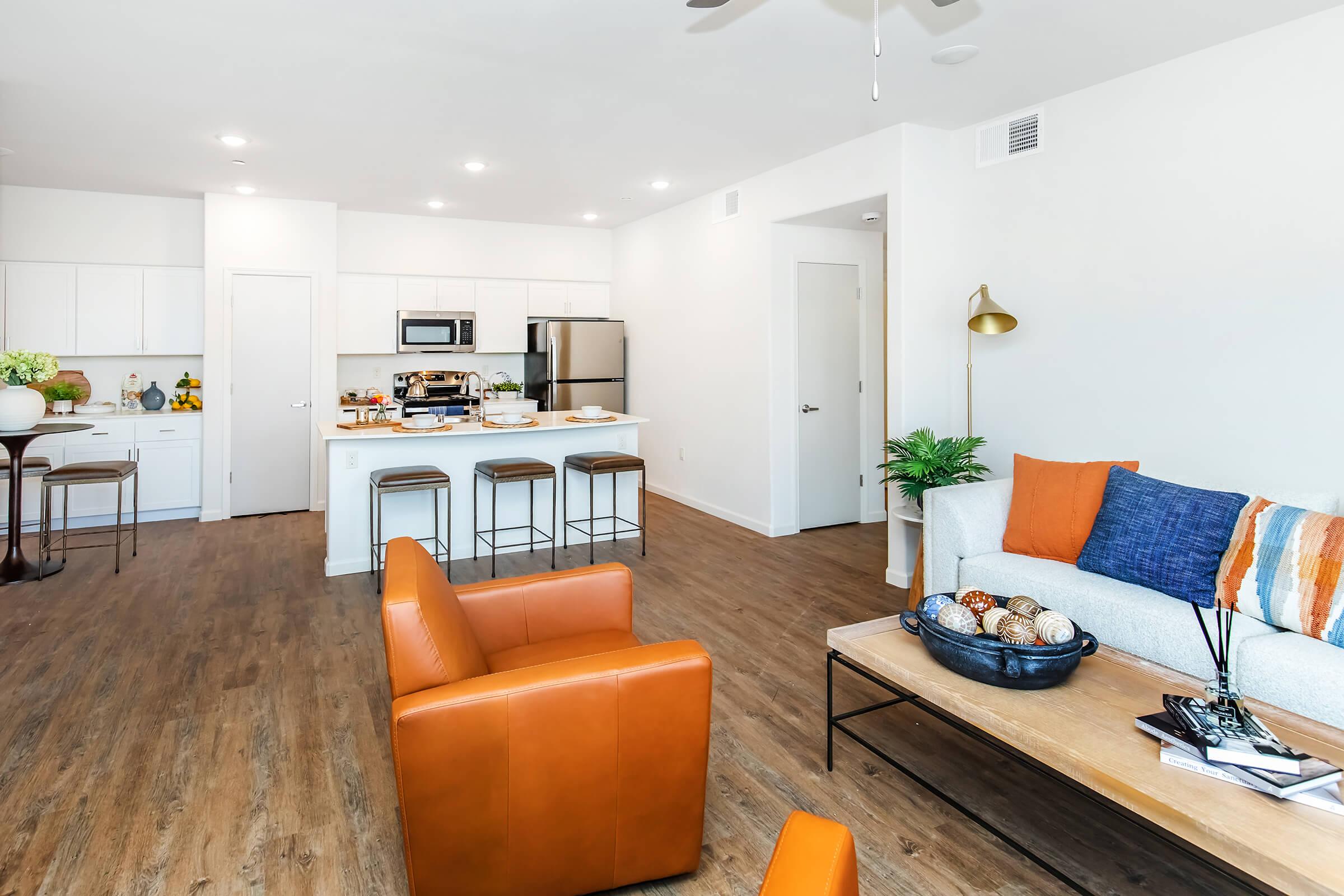 Modern open-concept living area featuring a kitchen with white cabinets and stainless-steel appliances, a cozy living room with an orange armchair and decorative table, and a stylish coffee table with decorative items. Natural light brightens the space, creating a warm and inviting atmosphere.