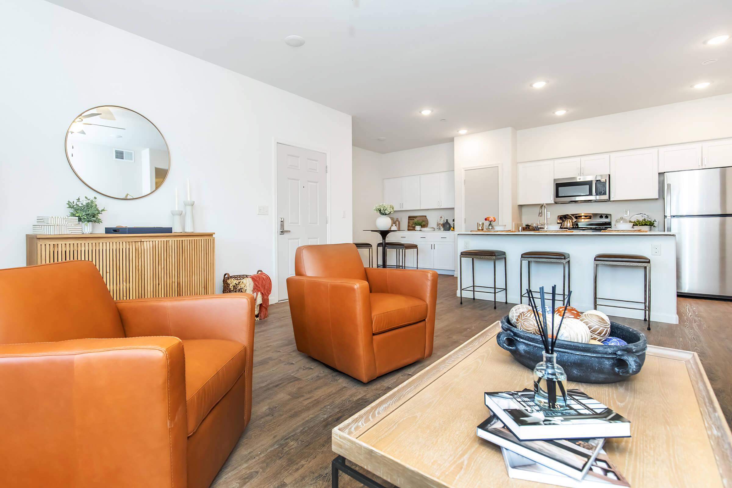A modern living space featuring two stylish orange leather armchairs and a wooden coffee table with decorative books. In the background, a contemporary kitchen is visible with stainless steel appliances and white cabinetry, complemented by a round wall mirror. The overall decor is bright and inviting.