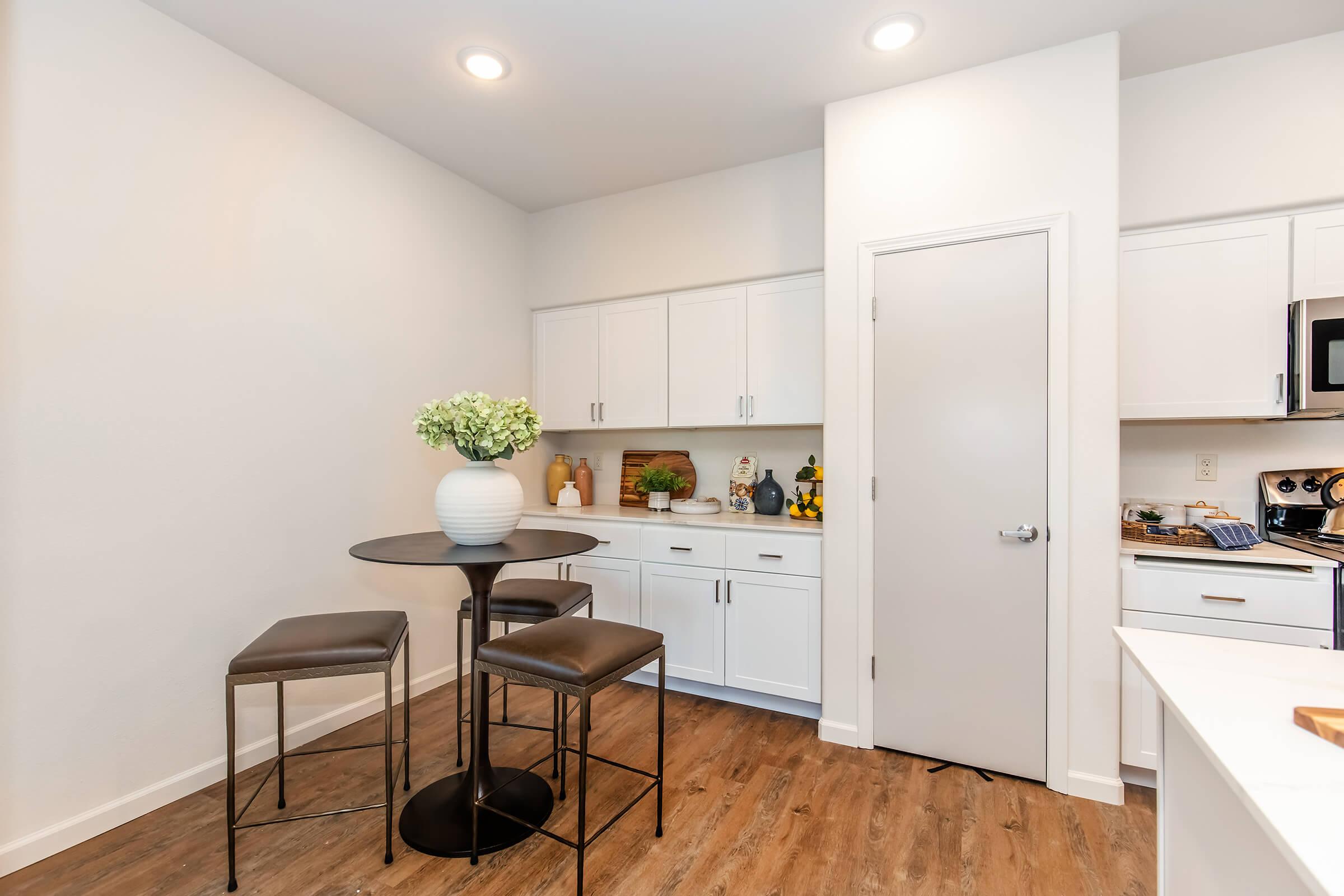Modern kitchen corner featuring a small round black table with two dark stools, a white vase with greenery on the table, and sleek white cabinets. The space has warm wood flooring and a light-colored wall, creating a bright and inviting atmosphere. A door leads to another area of the kitchen.