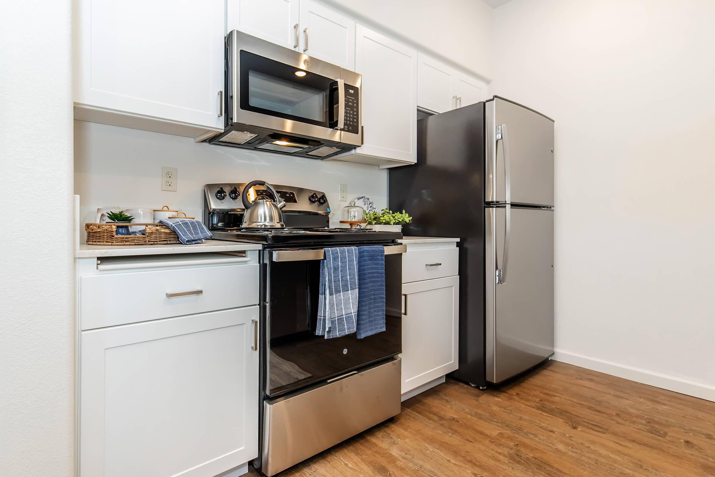 Modern kitchen featuring white cabinets, stainless steel appliances including a microwave, stovetop, and refrigerator. There is a kettle on the stove and a dish towel hanging near the oven. The countertop is neatly organized with a small plant and a basket, and the floor is wood laminate.