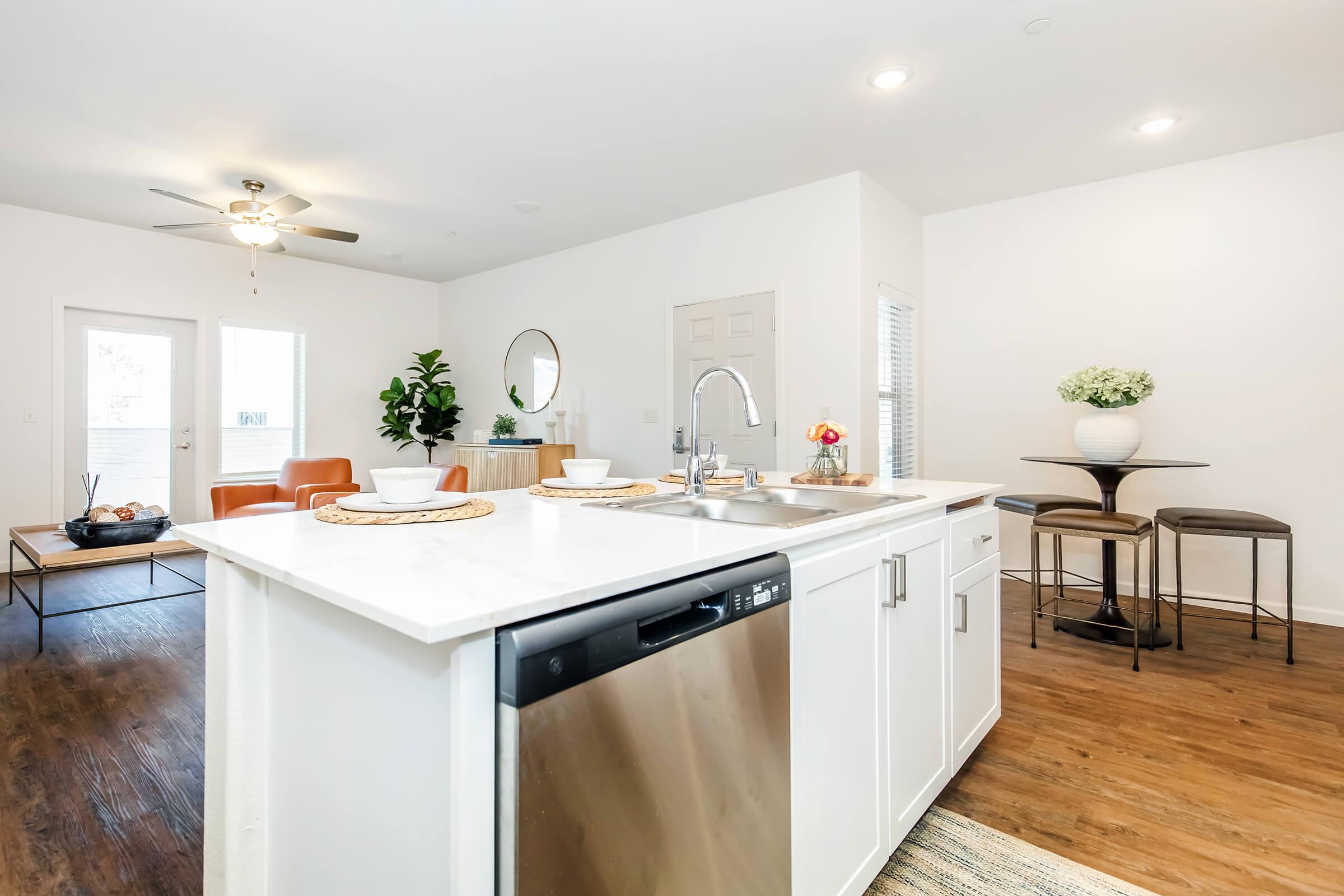 A modern kitchen with a white island featuring a sink and dishwasher. In the background, there's a cozy living area with orange chairs, a coffee table, and a plant. A small dining area has a round table with two stools. Natural light enters through windows, creating a bright and inviting atmosphere.