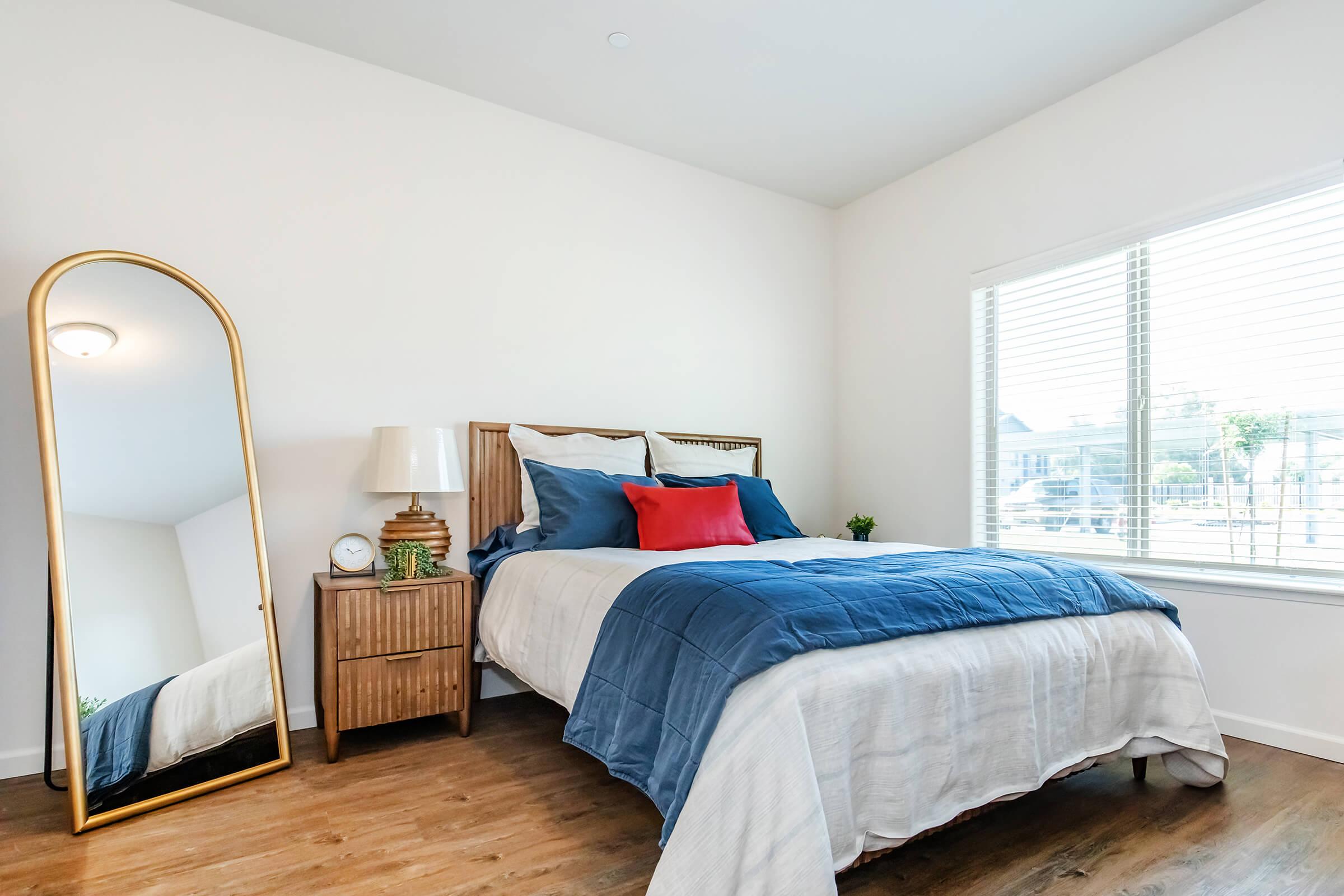 A cozy bedroom featuring a comfortable bed with blue and white bedding and a red decorative pillow. A tall, golden-framed mirror leans against the wall beside a wooden nightstand with a lamp and a small plant. Natural light streams in through a window adorned with blinds.