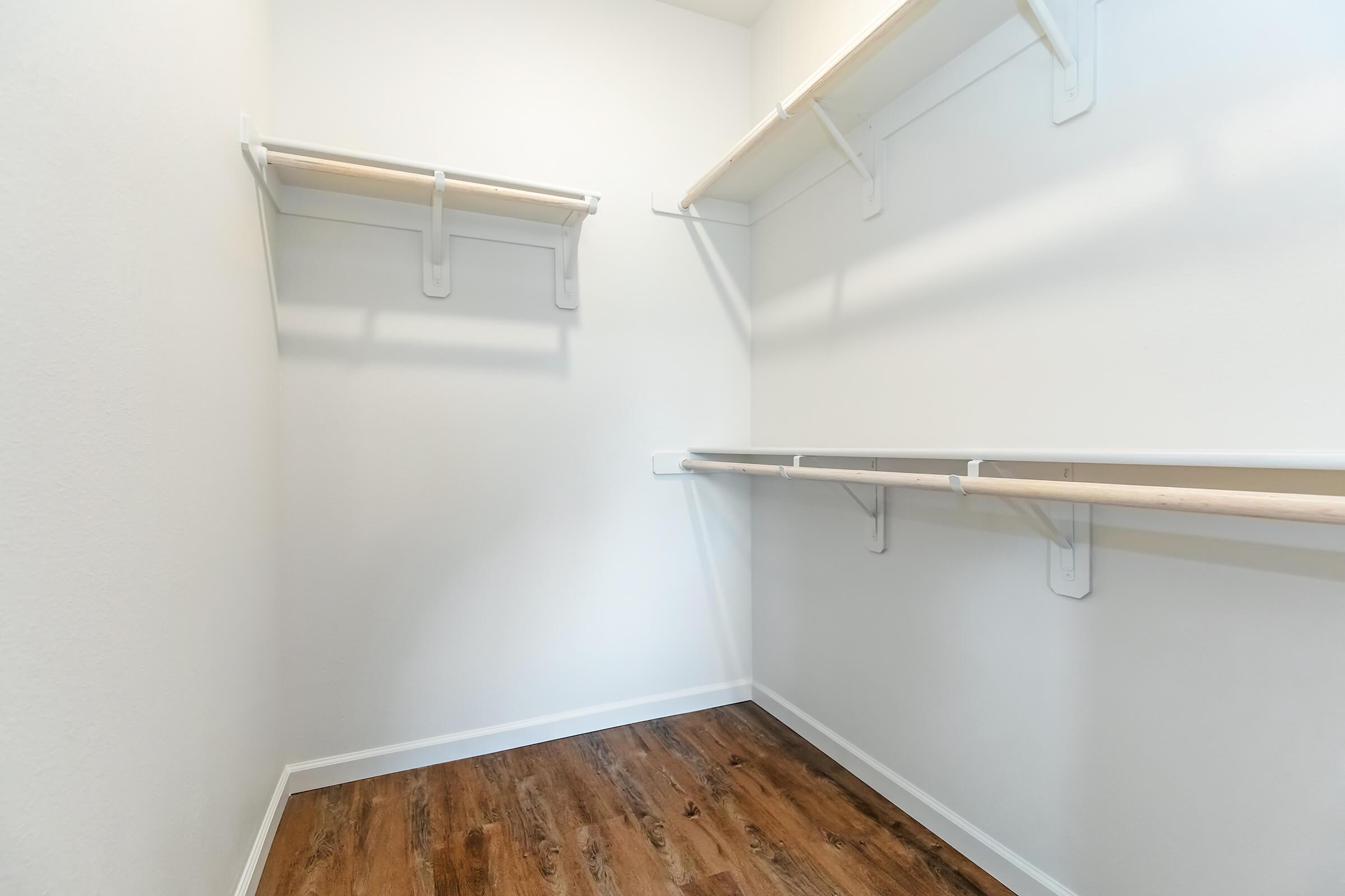 A spacious and empty closet featuring light-colored walls, wooden flooring, and multiple shelving units along the sides for storage, creating a clean and organized appearance.