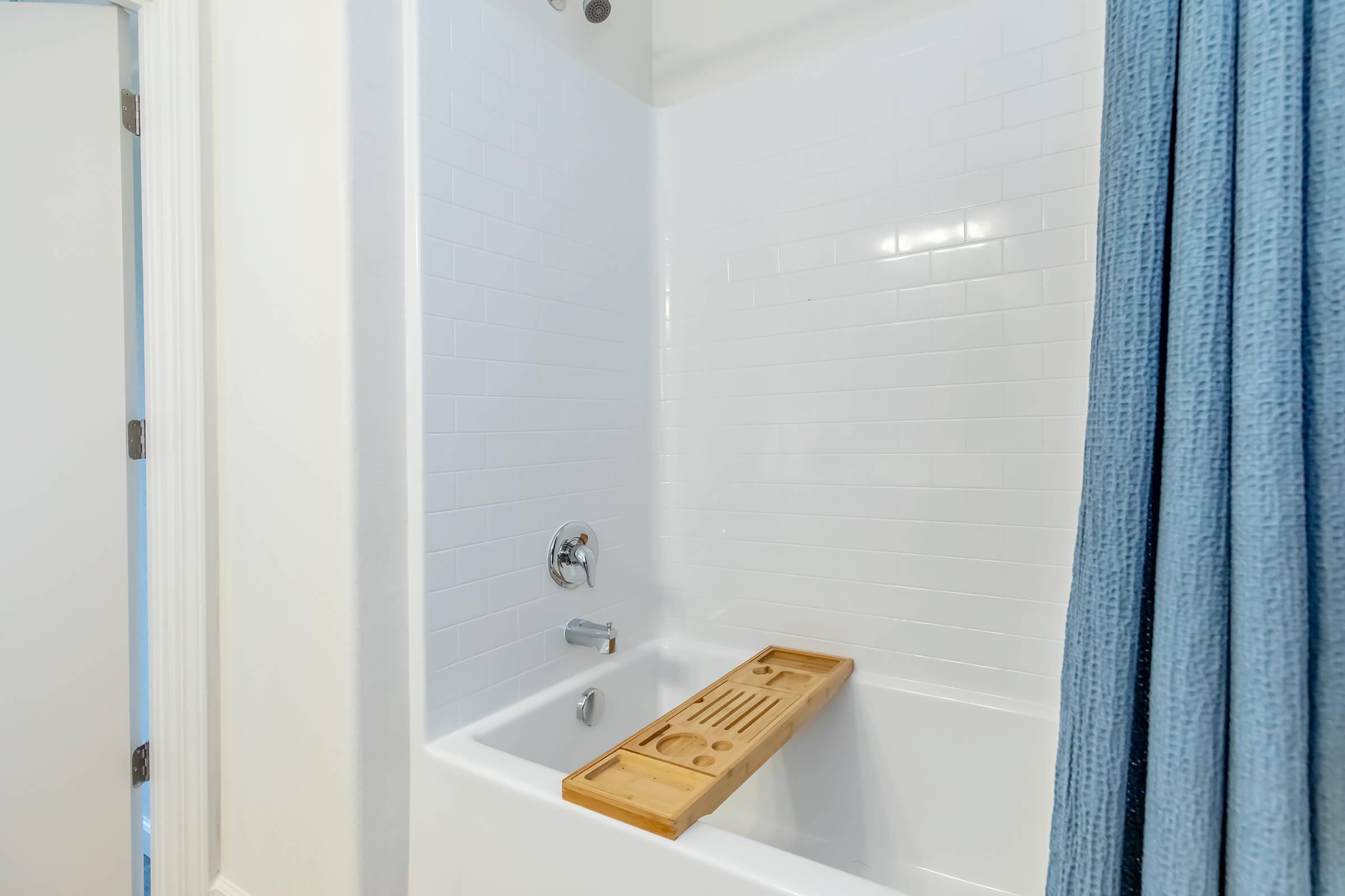 A clean and modern bathroom featuring a white tiled bathtub with a wooden bath tray placed across the top. The space is brightened by natural light, and there is a light blue shower curtain hanging nearby. The overall atmosphere is serene and inviting.