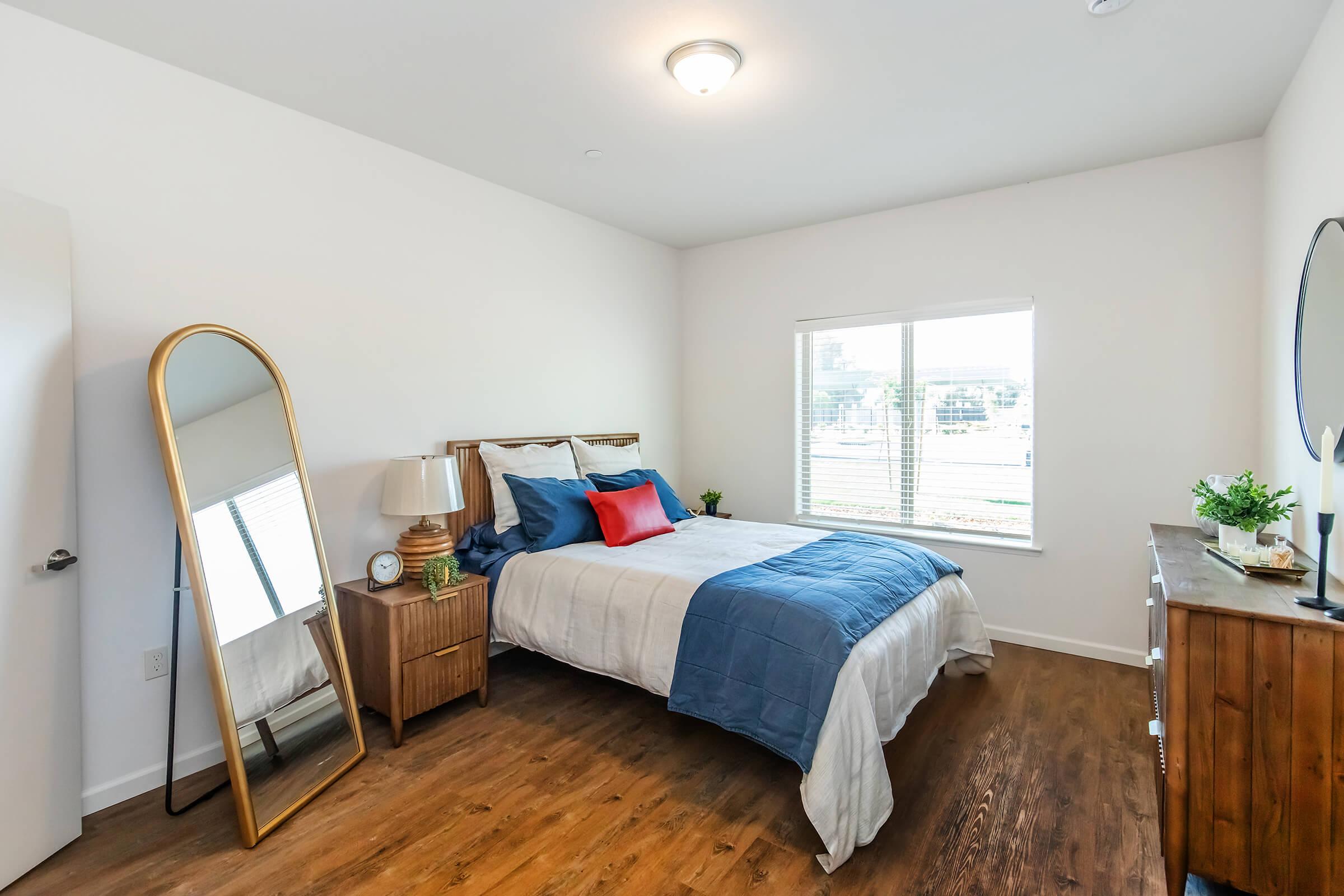 A well-lit, modern bedroom featuring a bed with blue and white bedding, a large mirror, a bedside lamp, and a wooden dresser. There is a window with light curtains, allowing natural light to fill the space, and decorative plants adding a touch of greenery. The floor is wooden, enhancing the room's cozy feel.