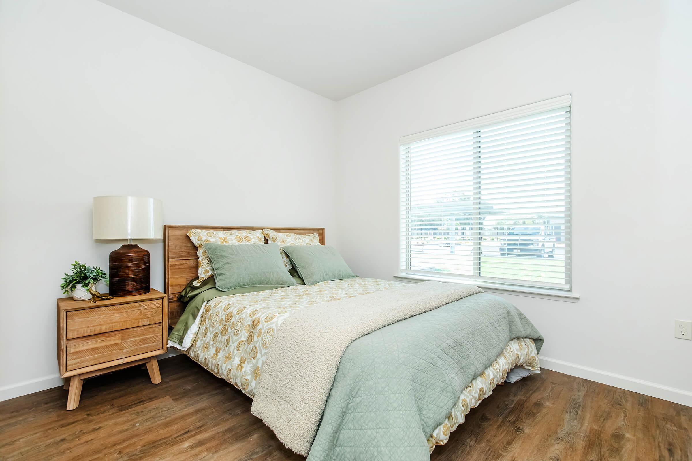 A cozy bedroom featuring a wooden bed frame with a light green and patterned bedding set. There is a bedside table with a brown lamp and a small plant. The room has a window with blinds, allowing natural light to enter, and a clean, modern aesthetic with wooden flooring.