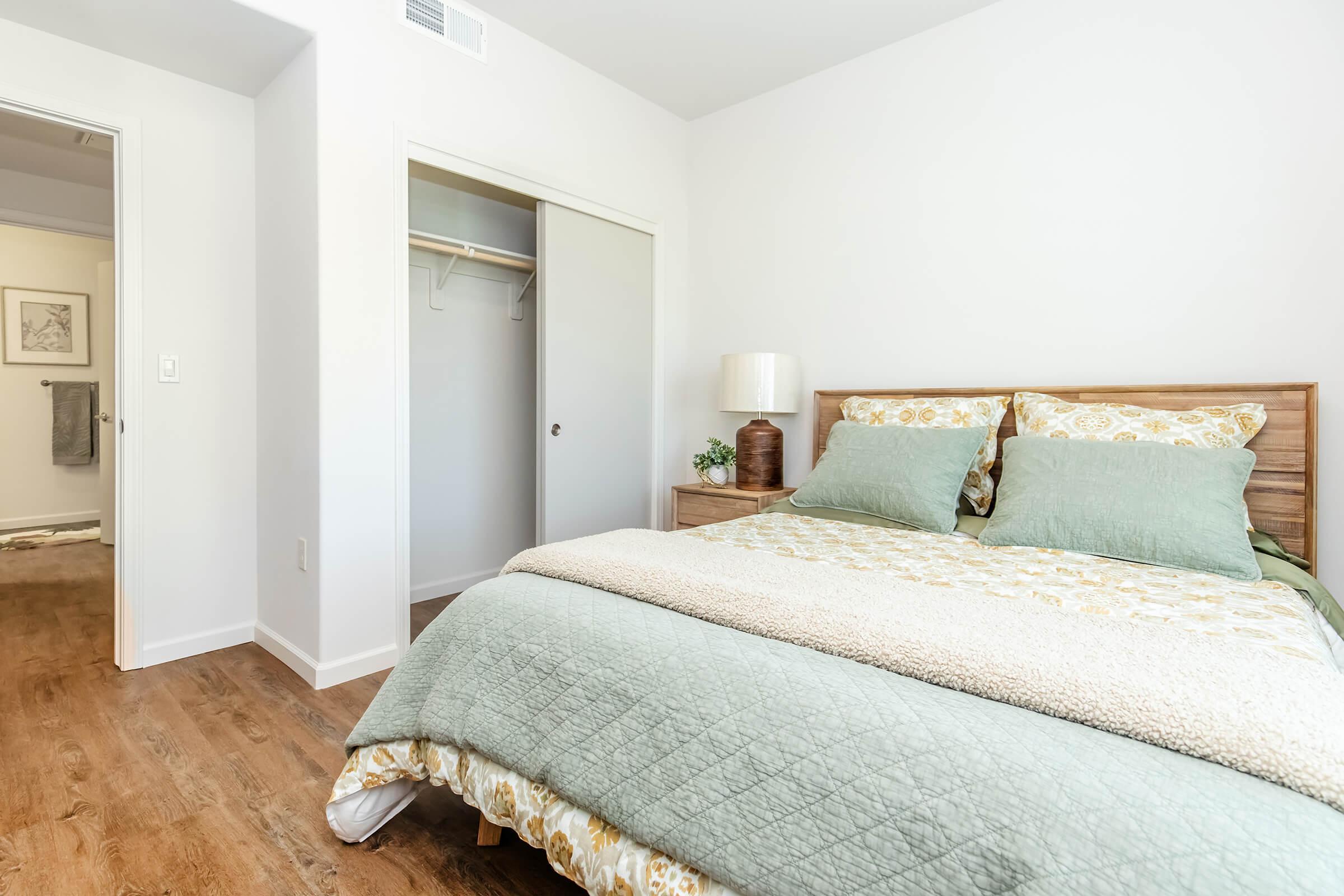 A cozy bedroom featuring a wooden bed with green and beige bedding. There's a bedside lamp and a small plant on the nightstand. In the background, an open closet door is visible, along with a doorway leading to another room. The walls are painted white, and the floor has wood-like flooring.