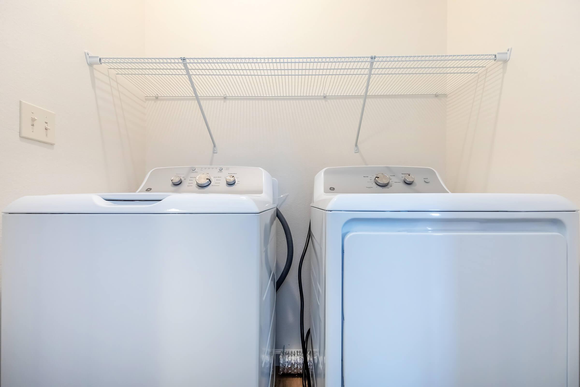 A side-by-side view of a white washing machine and dryer in a laundry room. The machines are placed against a wall, with a wire shelf mounted above them for storage. The setting is clean and organized, with a neutral wall color.