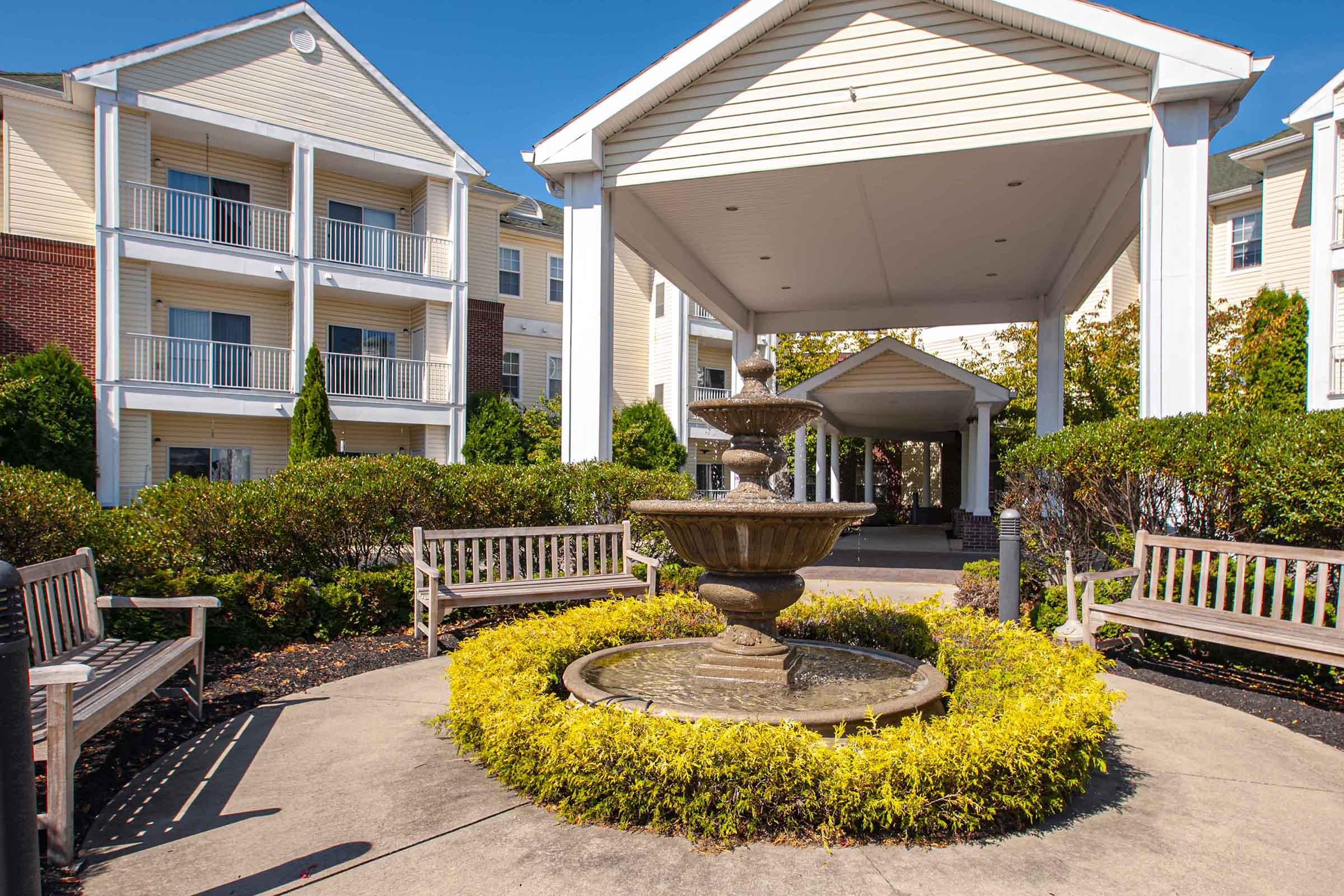 A peaceful courtyard featuring a stone fountain surrounded by neatly trimmed bushes and benches. The background includes a multi-story building with balconies, all under a clear blue sky. Sunlight creates a vibrant and inviting atmosphere.