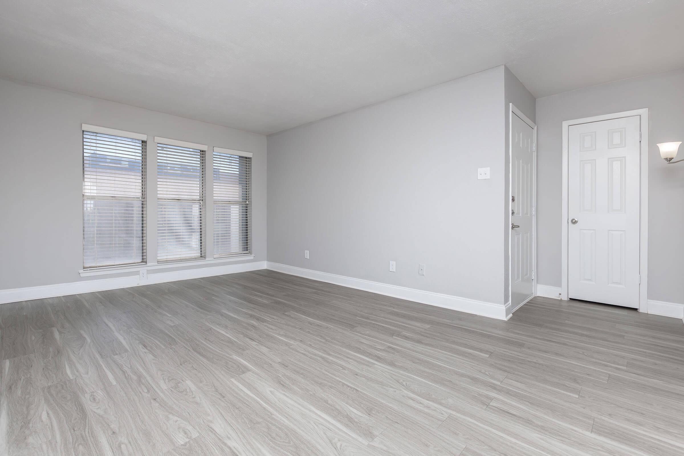 Empty room with light gray walls and light-colored vinyl flooring. Features include a set of three windows with blinds on one side and a white door leading to another space. The space is well-lit and has a clean, modern aesthetic.