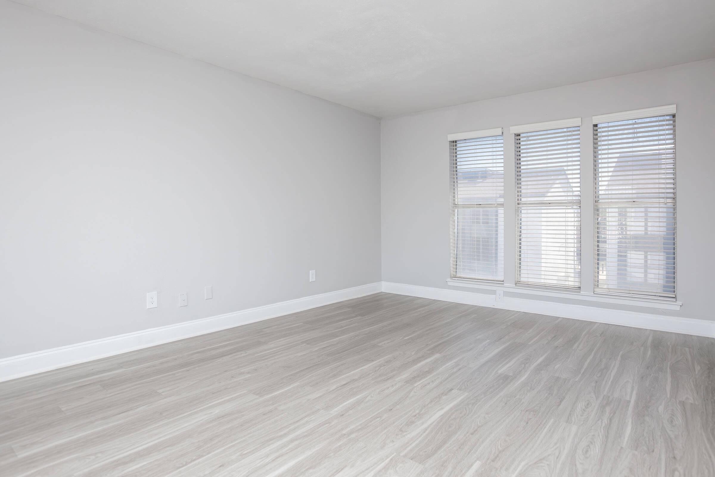 A spacious, empty room with light gray walls and a pale hardwood floor. It features three windows with blinds that allow natural light to fill the space, creating a bright and airy atmosphere. The room is free of furniture, showcasing its potential for various design options.