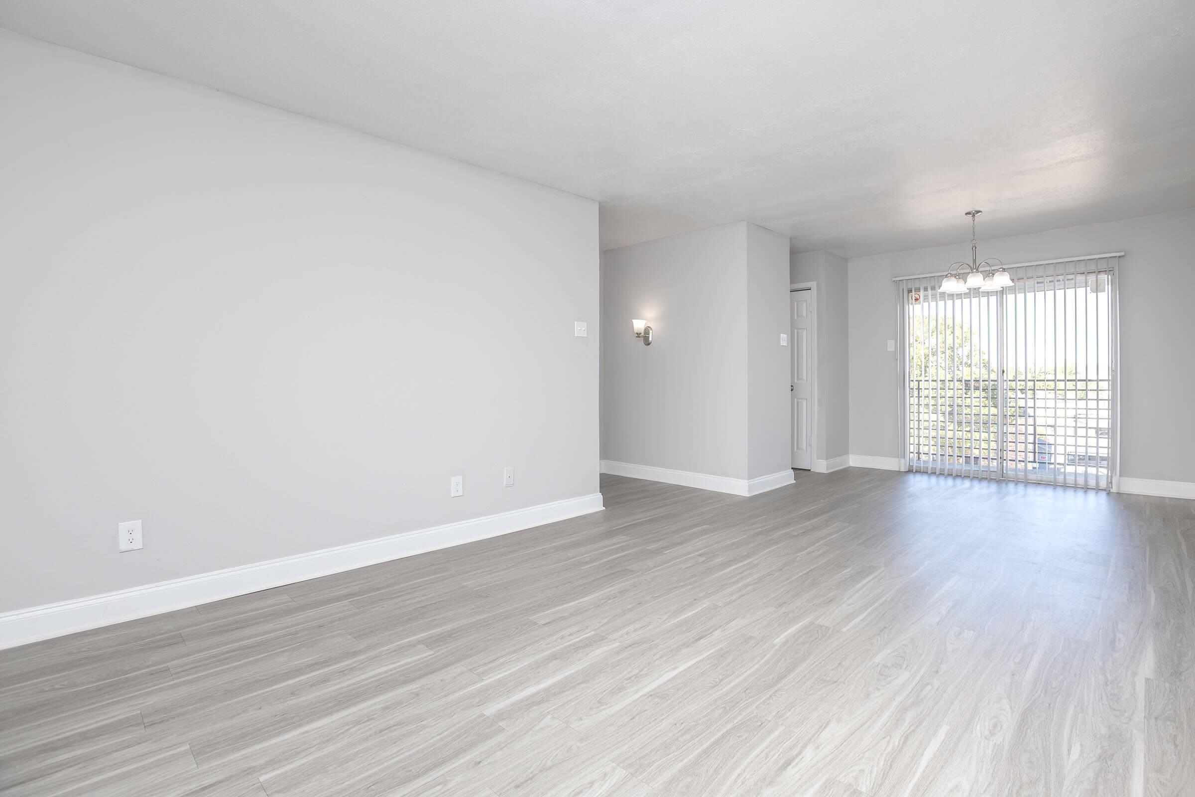 Spacious, empty room with light-colored wooden flooring and gray walls. A sliding door with vertical blinds leads to a balcony, allowing natural light to enter. Simple light fixtures are installed, and there is a door that likely leads to another room. The overall atmosphere is bright and modern.