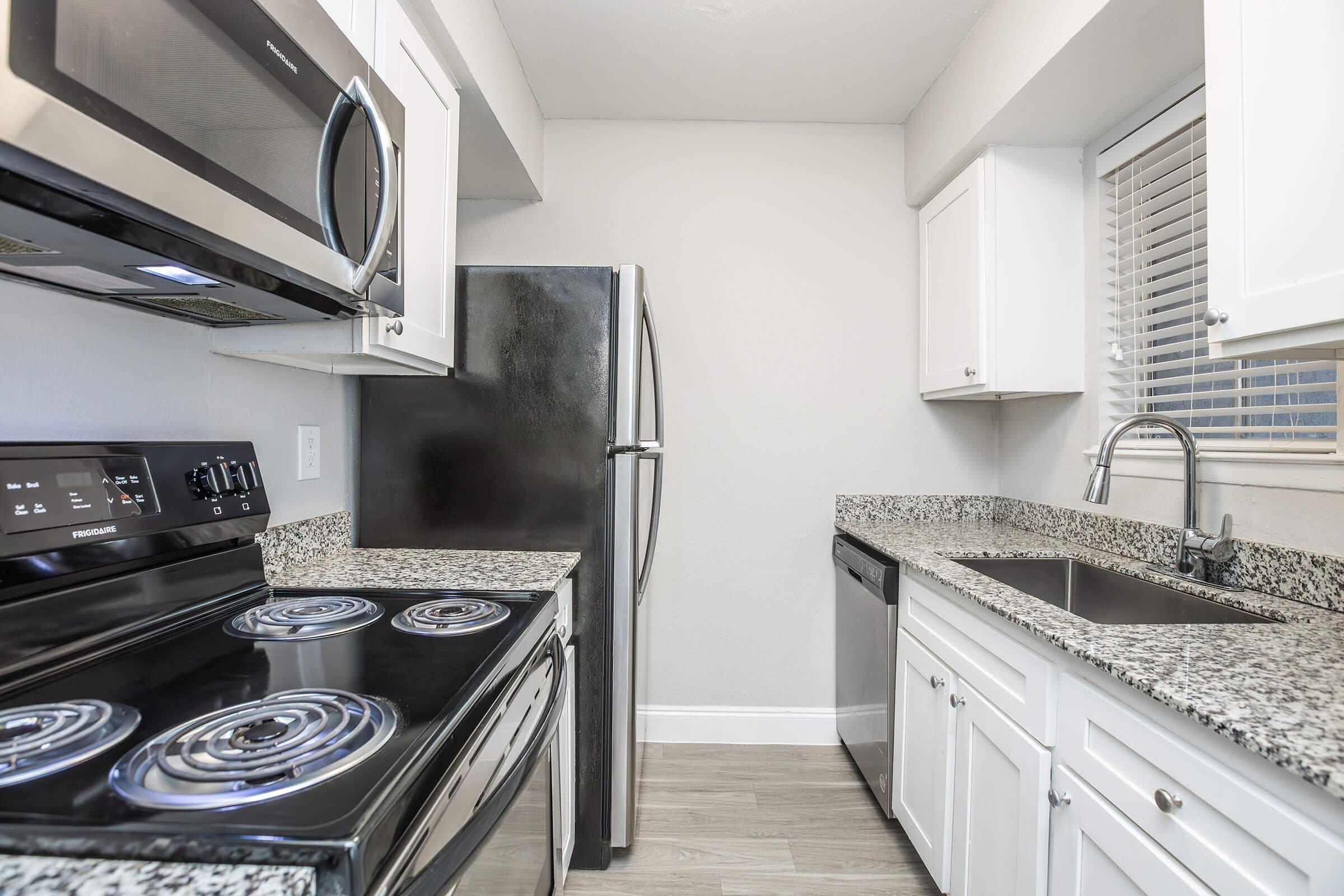 Modern kitchen featuring stainless steel appliances, including a microwave and refrigerator, black stove with an oven, and a dishwasher. The countertops are granite with a light gray finish, and the cabinets are white. Natural light enters through a window above the sink, enhancing the clean, contemporary look.