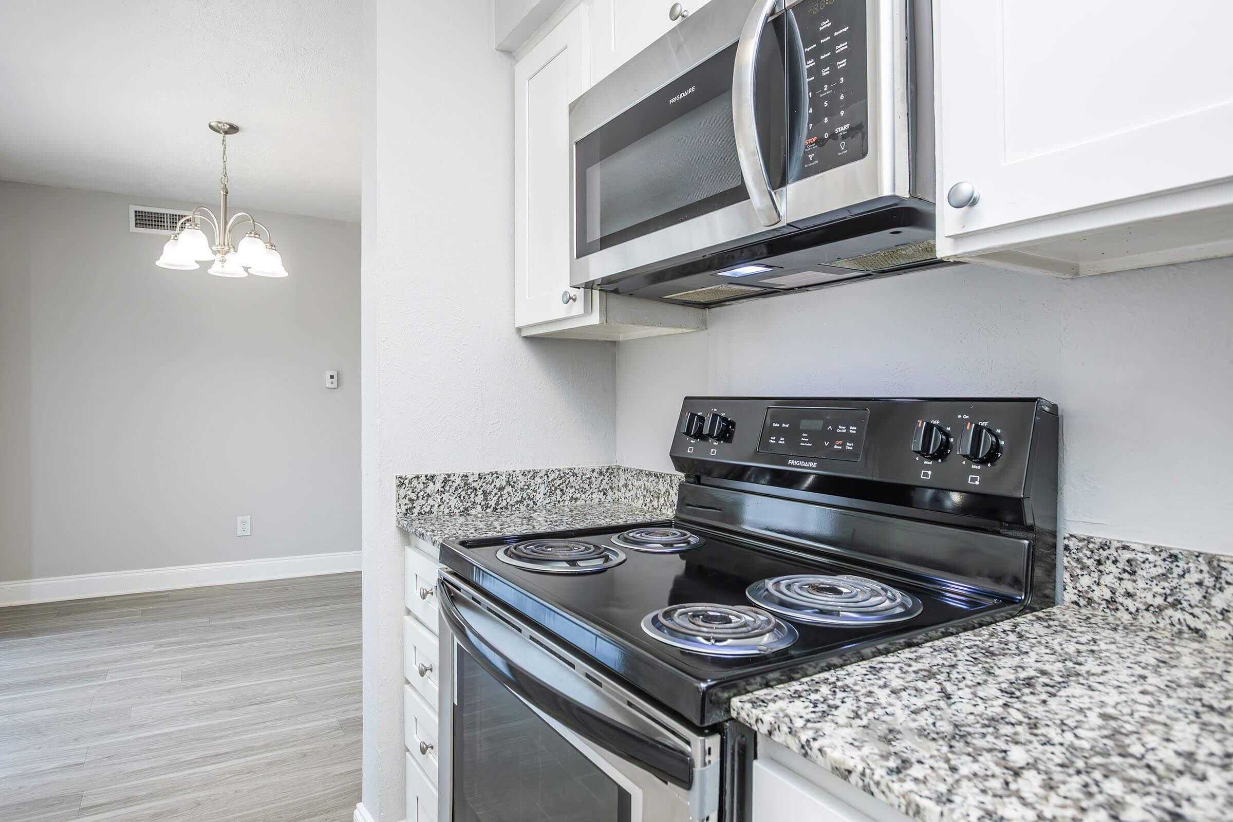 A modern kitchen featuring a black electric stove, a microwave above it, and granite countertops. White cabinets and light-colored walls create a bright and open atmosphere. In the background, a dining area with a light fixture is partially visible.