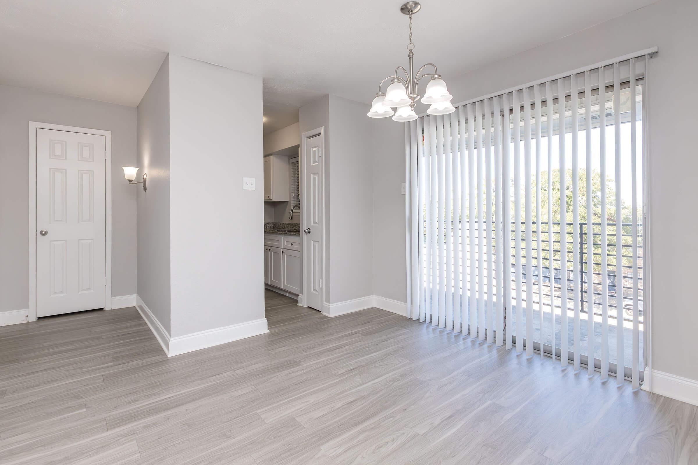 An interior view of a modern, light-colored room featuring a dining area, a sliding glass door with vertical blinds leading to a balcony, and a kitchen entrance on the left. The walls are painted in a soft gray, with a chandelier hanging from the ceiling, and a decorative wall sconce beside the door.
