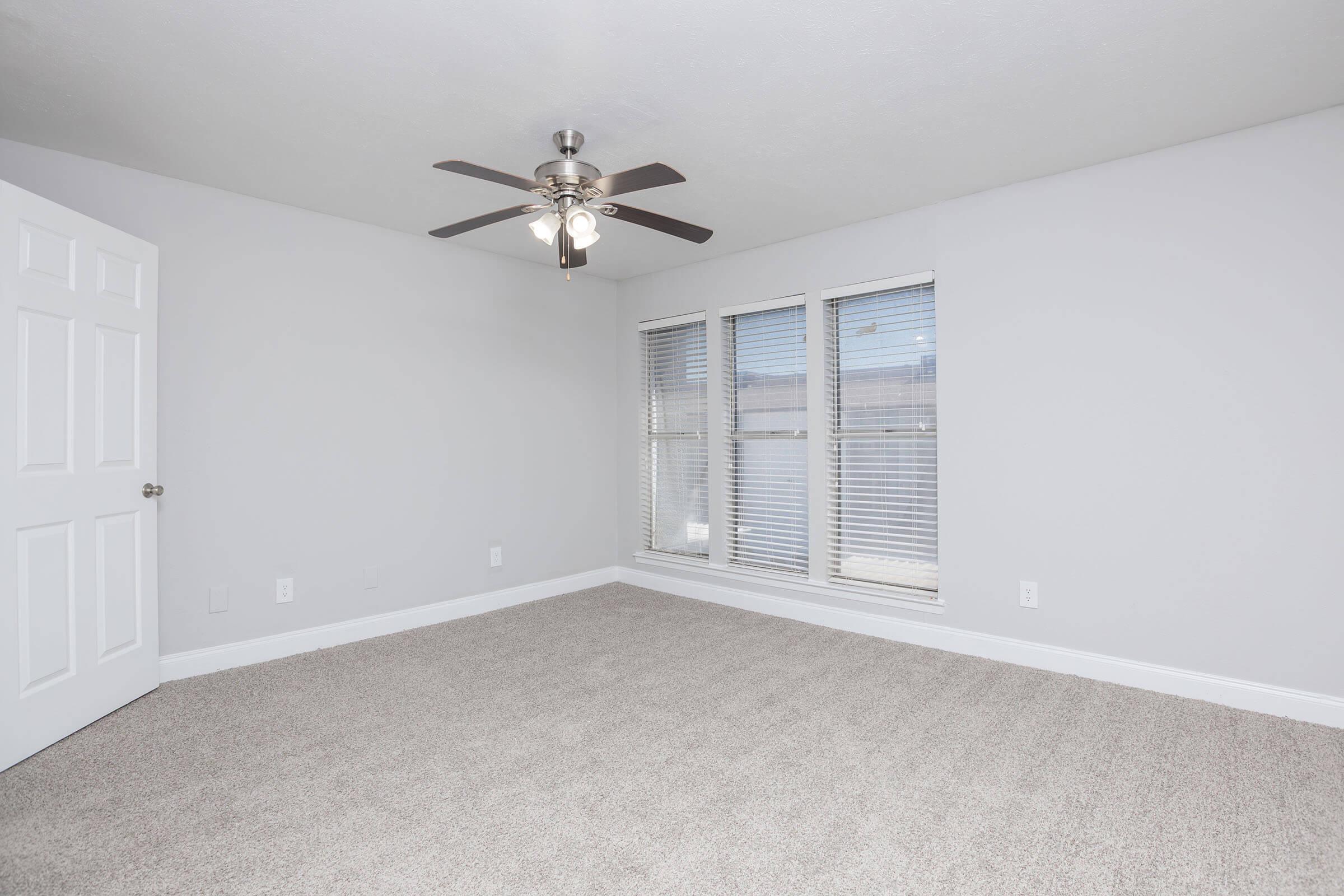 A spacious, empty room with light gray walls and new carpets. It features a ceiling fan with five blades, and large windows with blinds that allow natural light to fill the space. The door on the left is wooden and white, adding to the room's modern aesthetic.